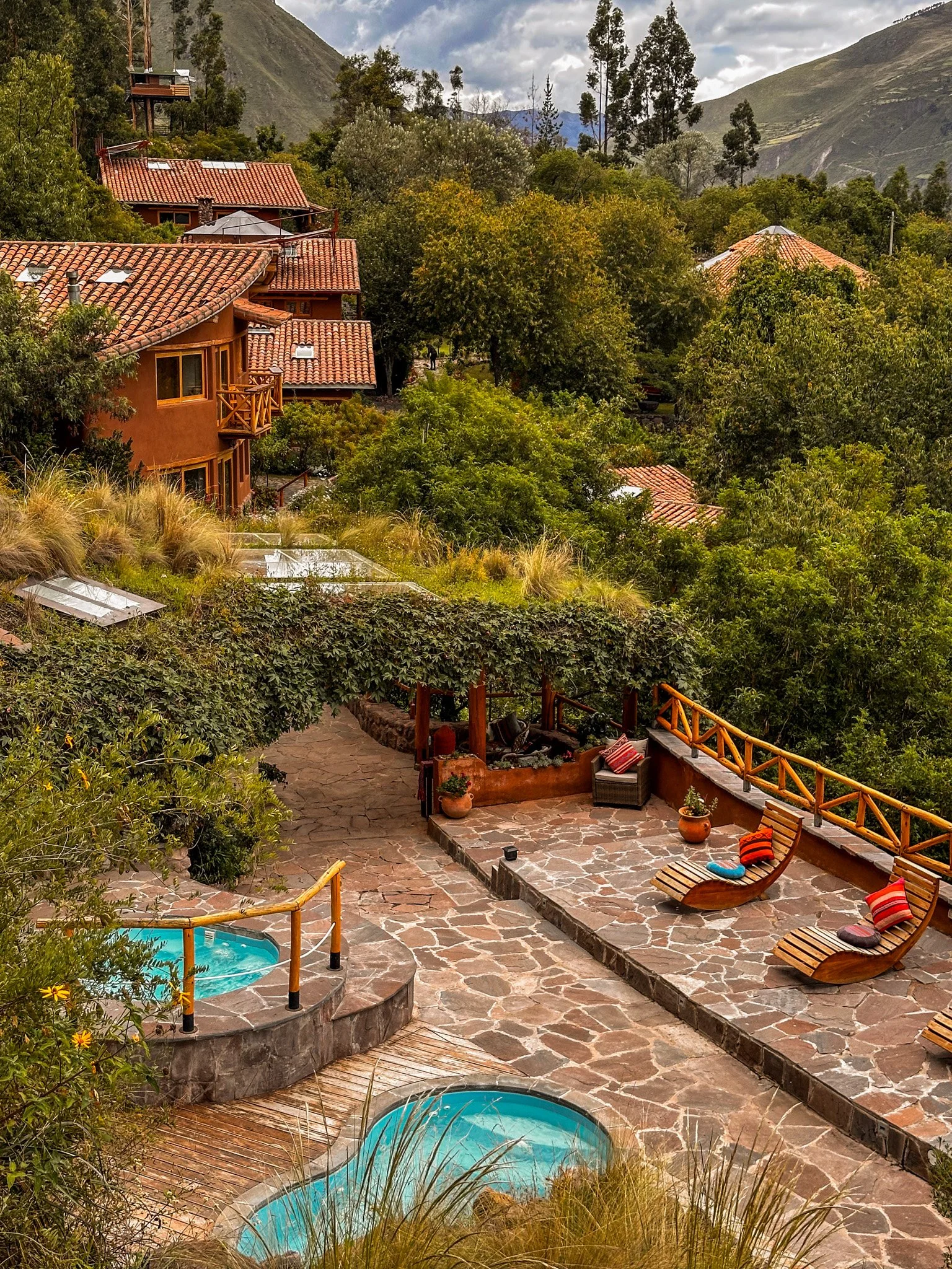 Outdoor terrace with stone flooring, two wooden lounge chairs with colorful cushions, small water pools, surrounded by lush green trees and mountain scenery in the background.