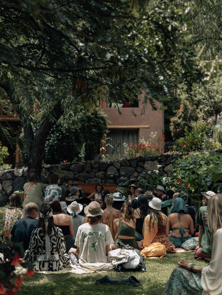 Group of people sitting outdoors on grass, some wearing summer clothes and hats, gathered under a large tree in a park or garden during daytime.