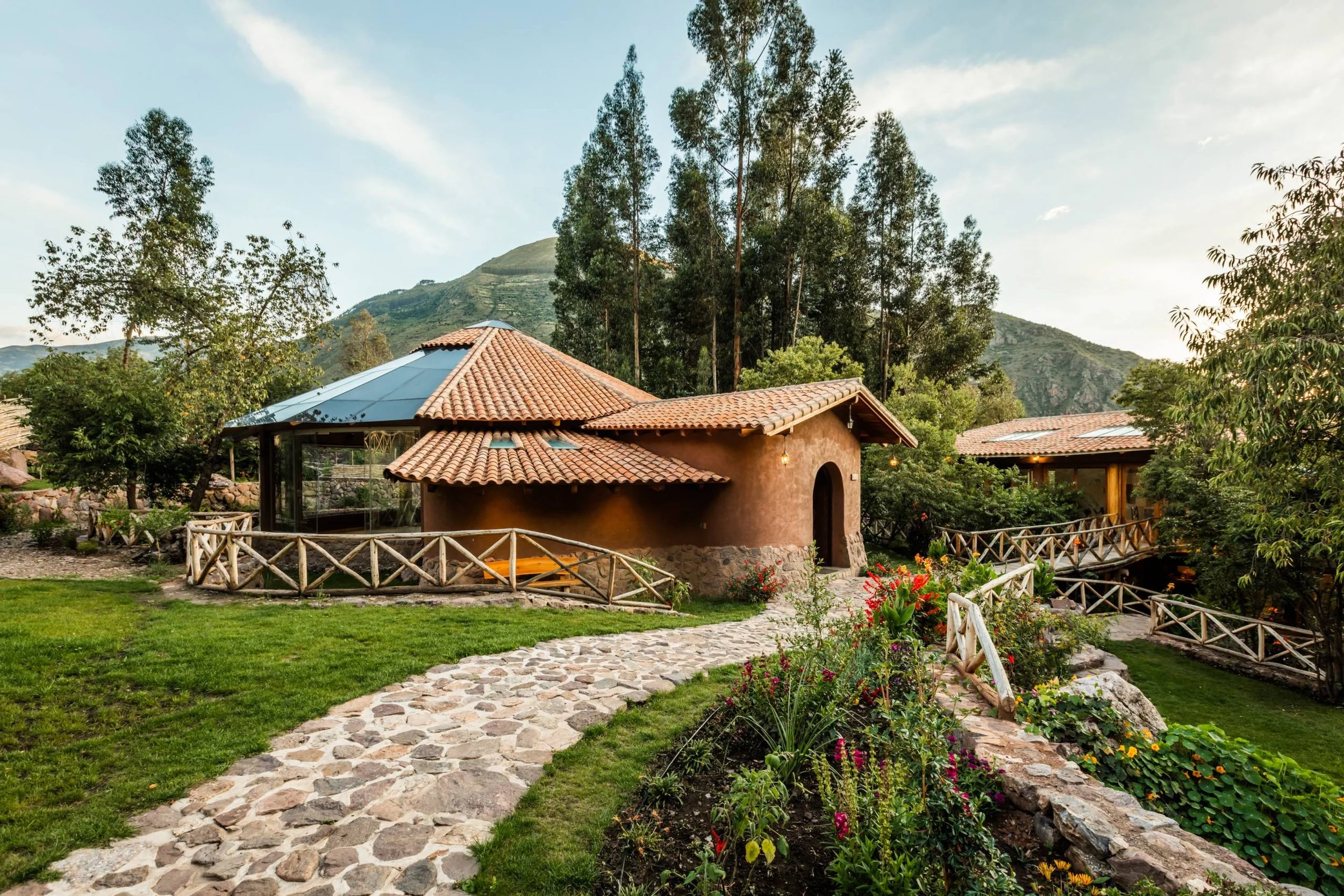 A scenic view of a house with a tiled roof, surrounded by a lush garden with flowers and trees, with mountains in the background and a blue sky above.