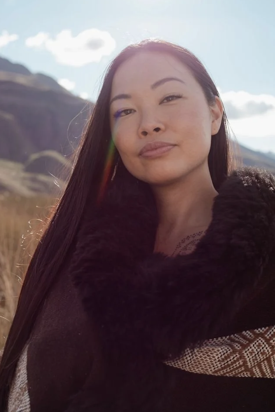 Tara Tao Center of Destiny with long black hair taking a selfie outdoors in a mountainous area during daytime, with sunlight and partly cloudy sky.