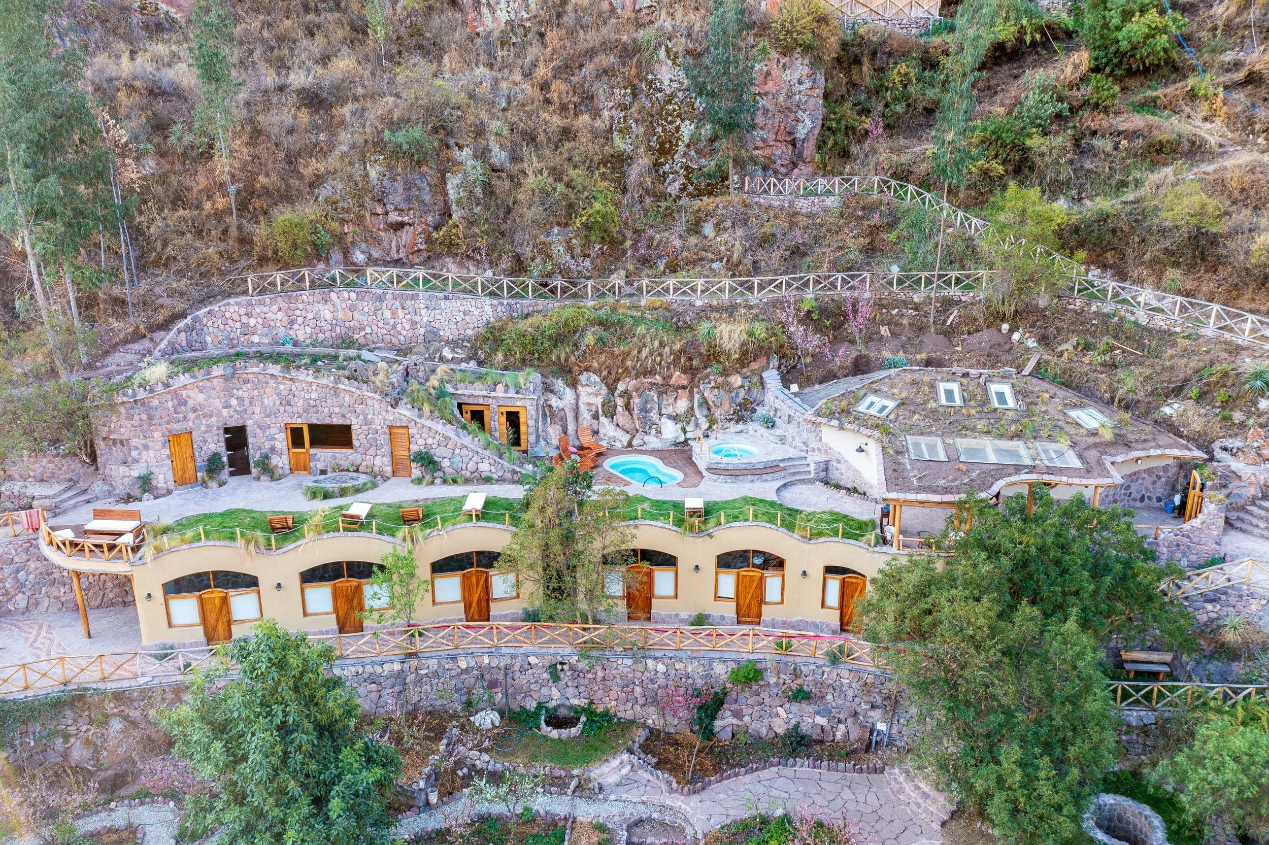 Aerial view of a hillside house with multiple levels, a swimming pool, patio furniture, and lush vegetation surrounded by a rocky landscape.