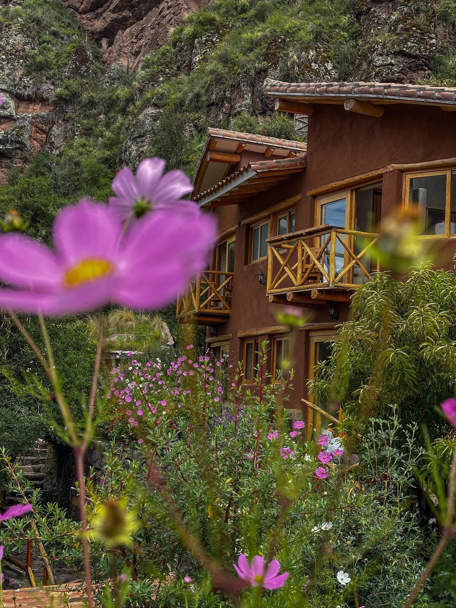 A house with a reddish-brown stucco exterior and wooden railings on a small balcony, surrounded by lush green plants and pink flowers in a garden, with a rocky hillside in the background.