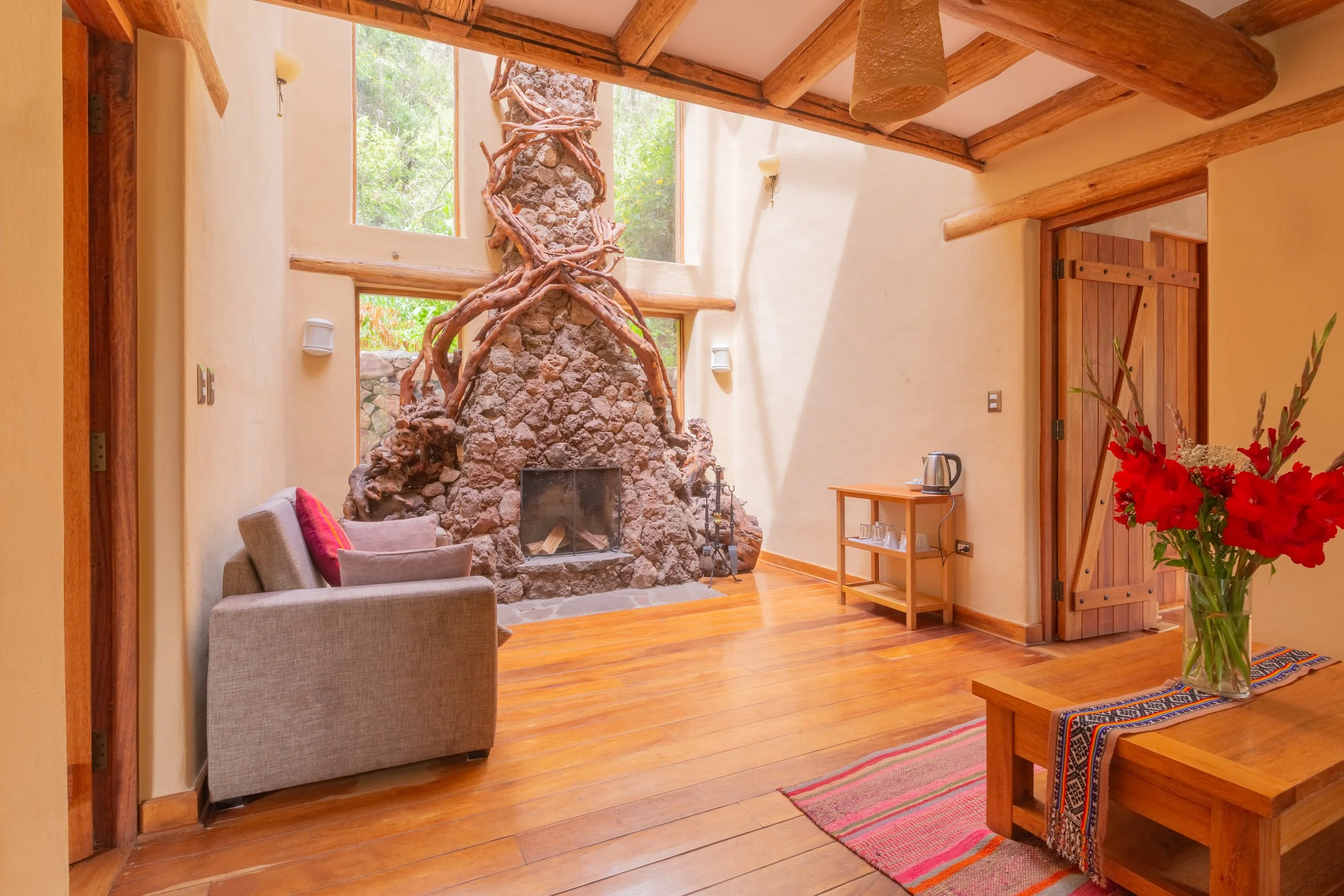 Living room with high wooden ceiling, large stone fireplace with driftwood, beige walls, wooden flooring, and a small wooden table with a silver electric kettle. There's a beige chair with a red pillow, a bouquet of red flowers, and a doorway with a wooden door.