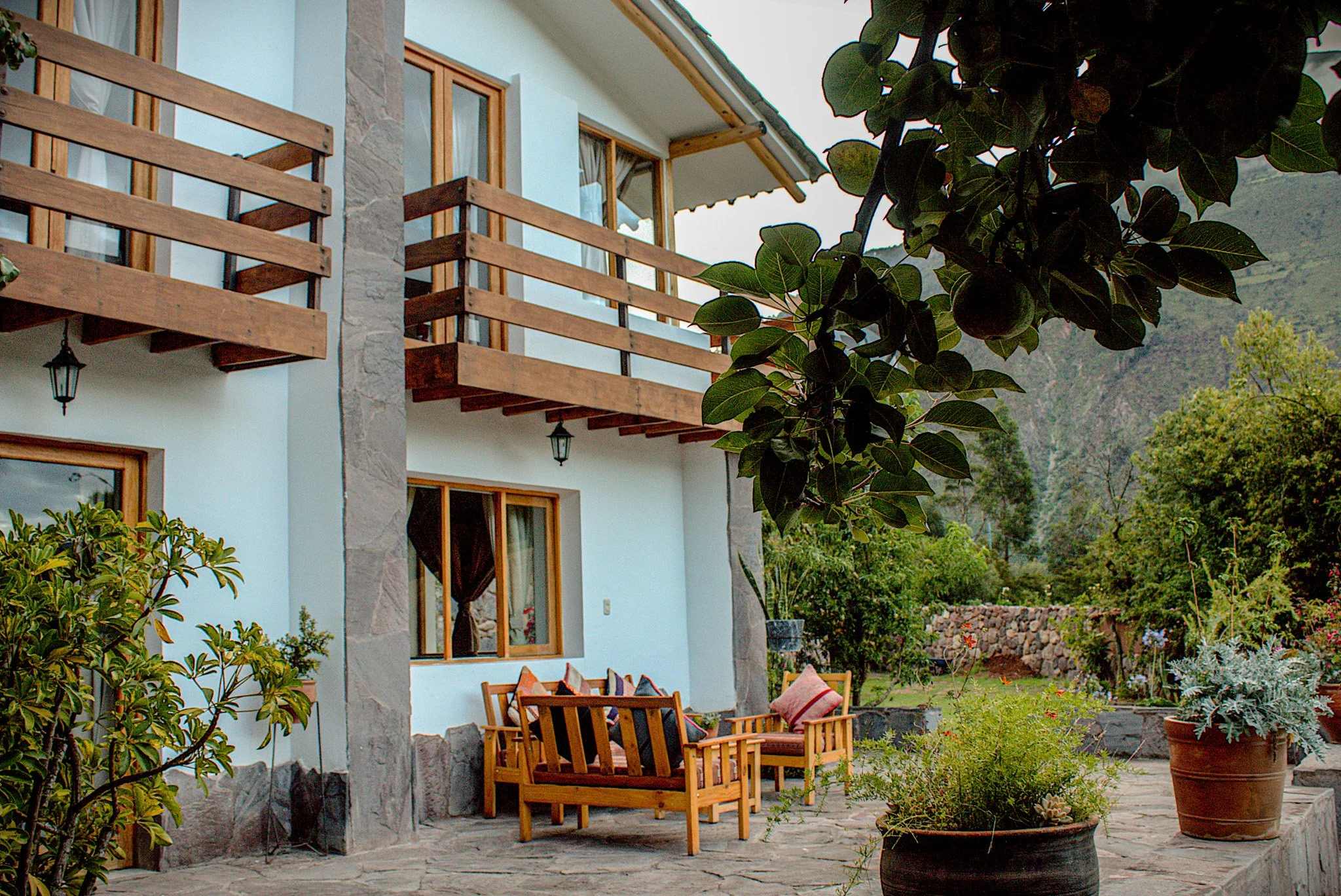 A balcony with wooden railing and exterior decor of a house, surrounded by green plants and potted flowers in a garden area, with mountains in the background.