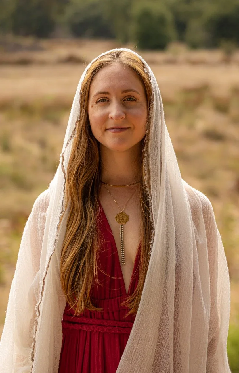 A young woman with long, wavy red hair standing outdoors in a natural setting, wearing a cream-colored shawl over a red dress, with layered gold necklaces.