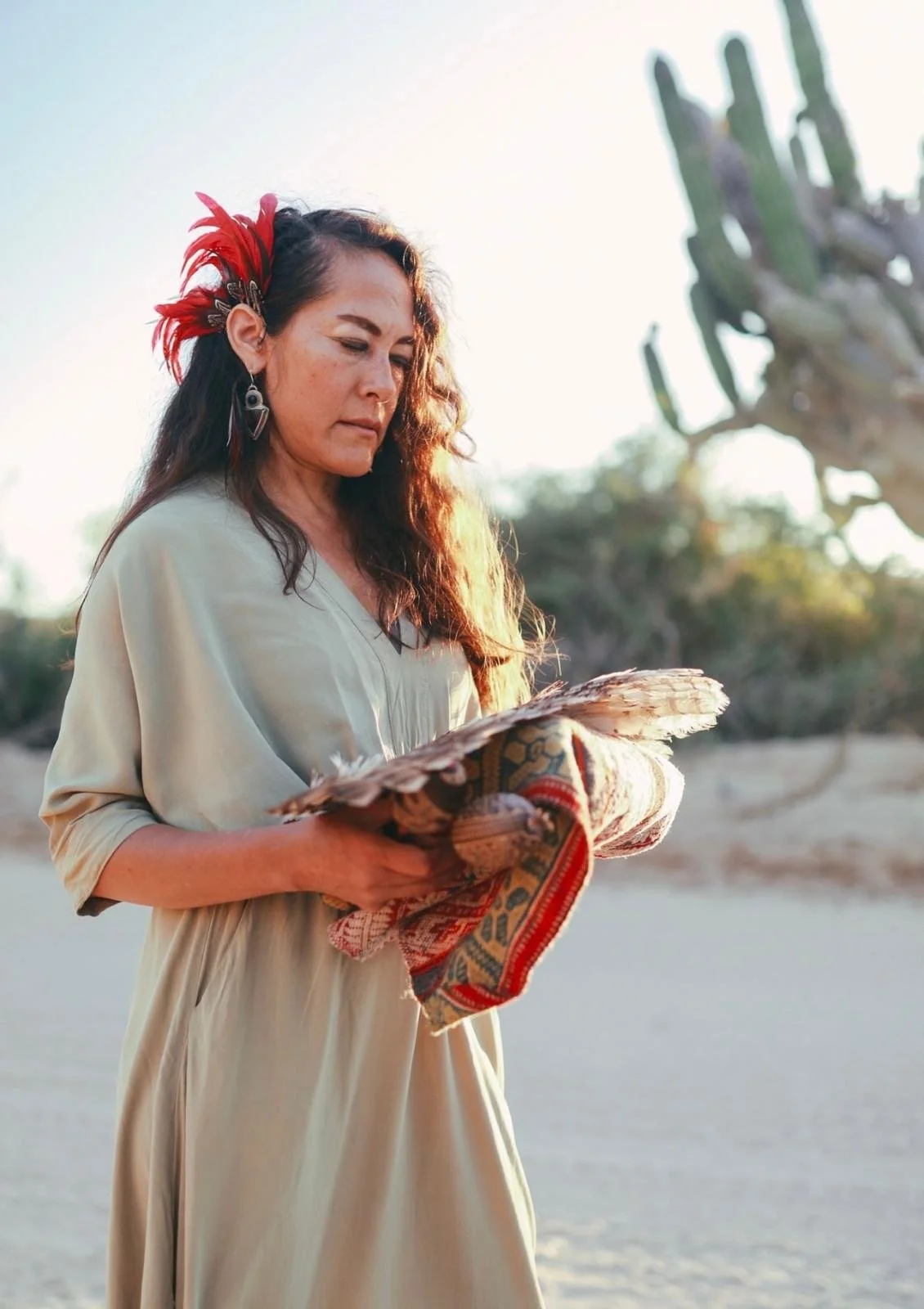 Woman with long hair, wearing earrings and a beige dress, holding a decorated feathered fan, standing outdoors near cactus plants during sunset.
