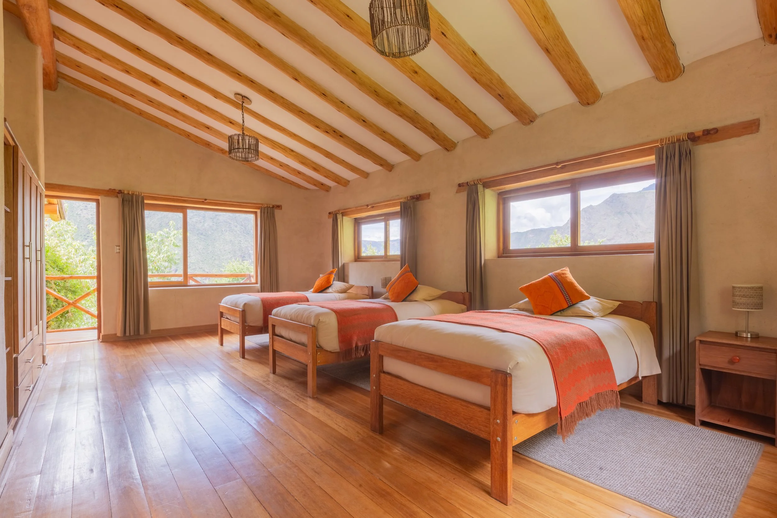 A cozy bedroom with three single beds, orange throw blankets, and matching orange pillows, wooden floors, large windows with mountain views, beige walls, and rustic wooden ceiling beams.