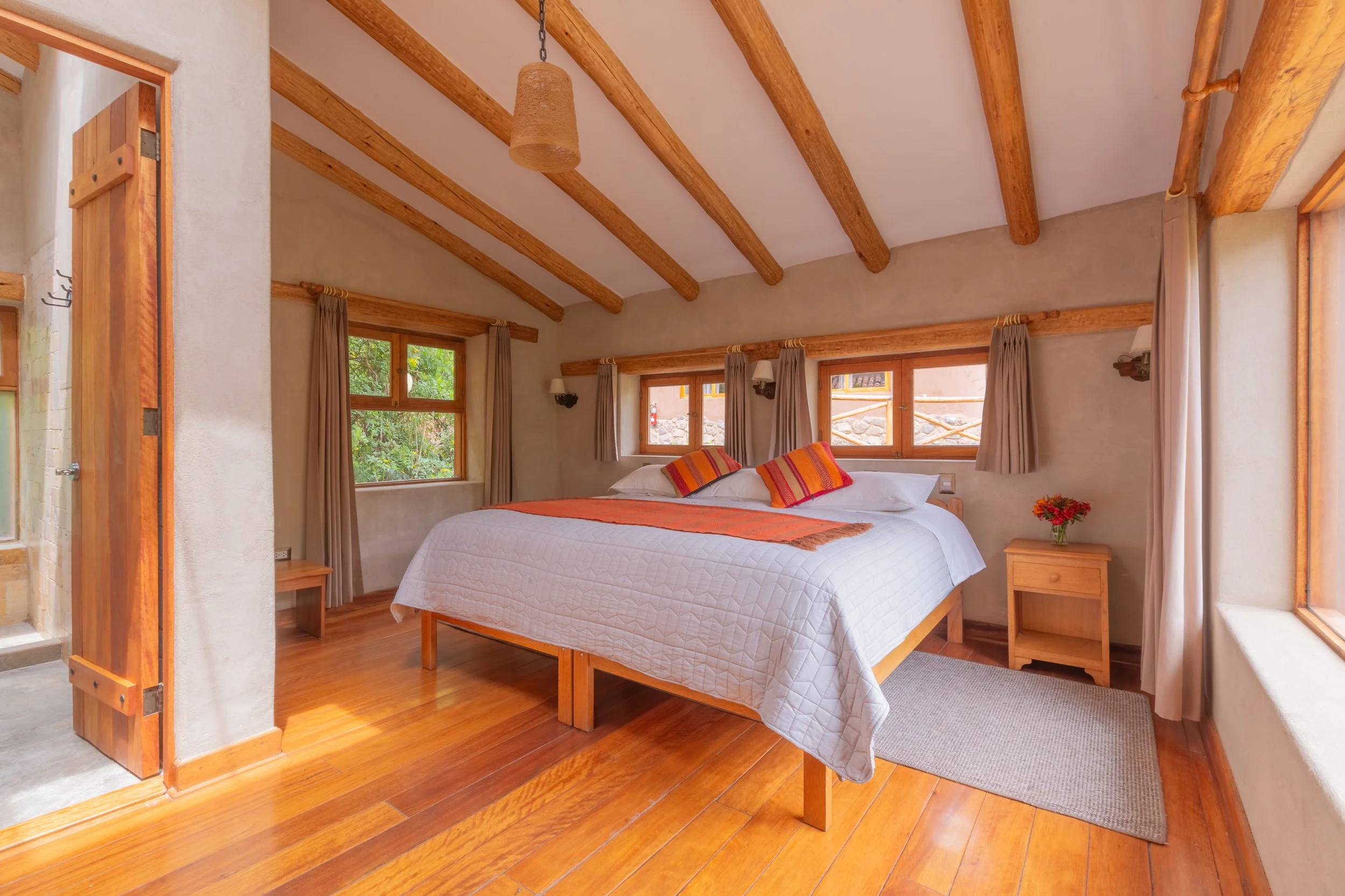 A cozy bedroom with a wooden bed frame, white quilt, and colorful pillows. The room has large windows with curtains, wooden ceiling beams, and a hanging light fixture.