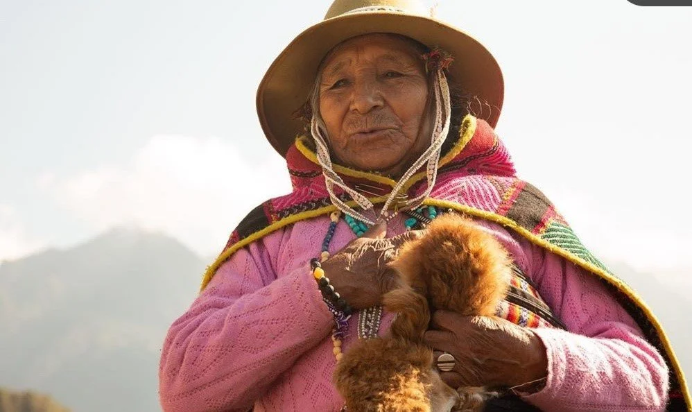 Elderly woman wearing traditional colorful clothing and a wide-brimmed hat, holding a small brown dog.