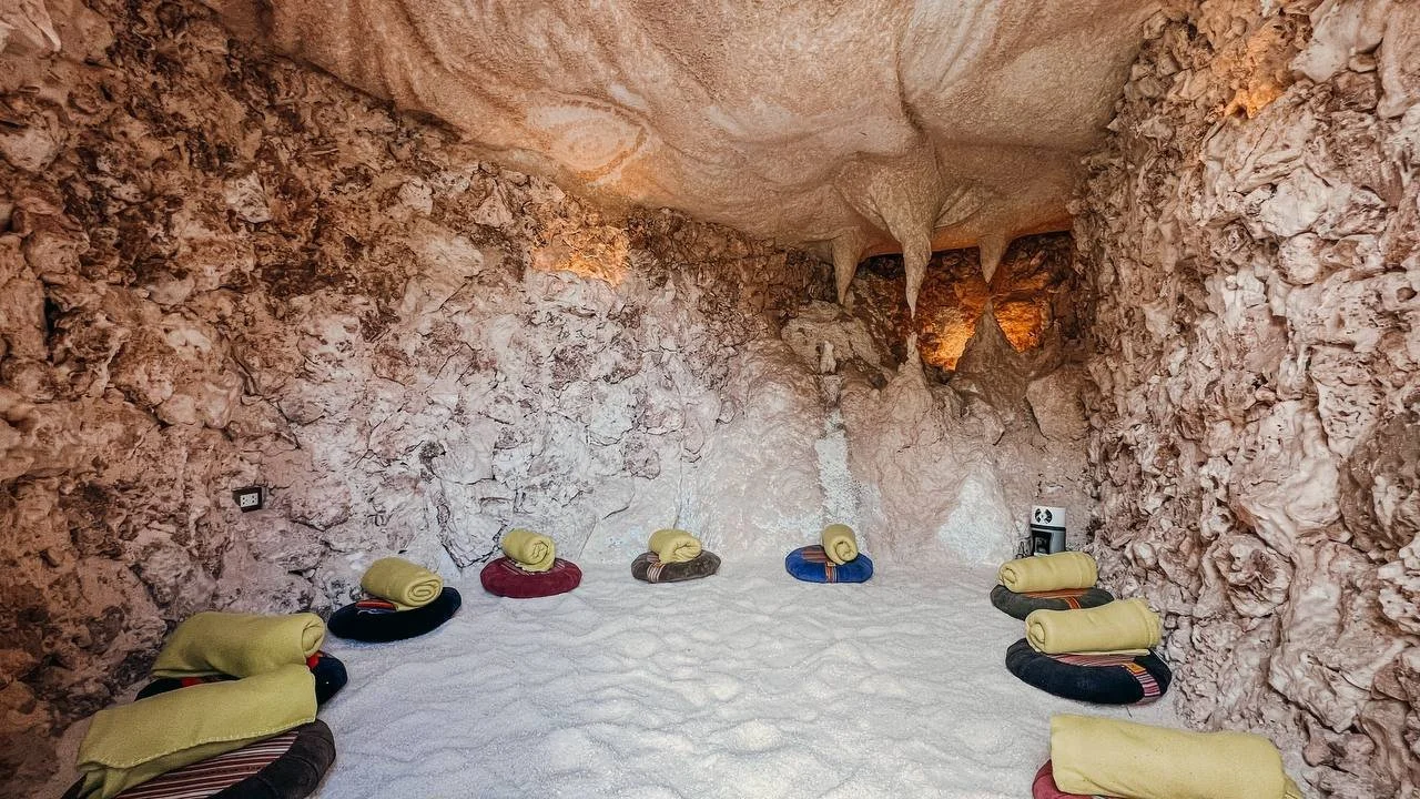 Interior of a salt cave with cushioned mats and yellow blankets arranged on the salt floor, and rough salt walls with stalactites hanging from the ceiling.