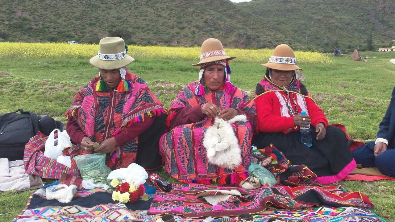 Three indigenous women in traditional colorful clothing and hats sit on a blanket on the grass, surrounded by offerings, flowers, and handmade crafts, with a field and mountains in the background.
