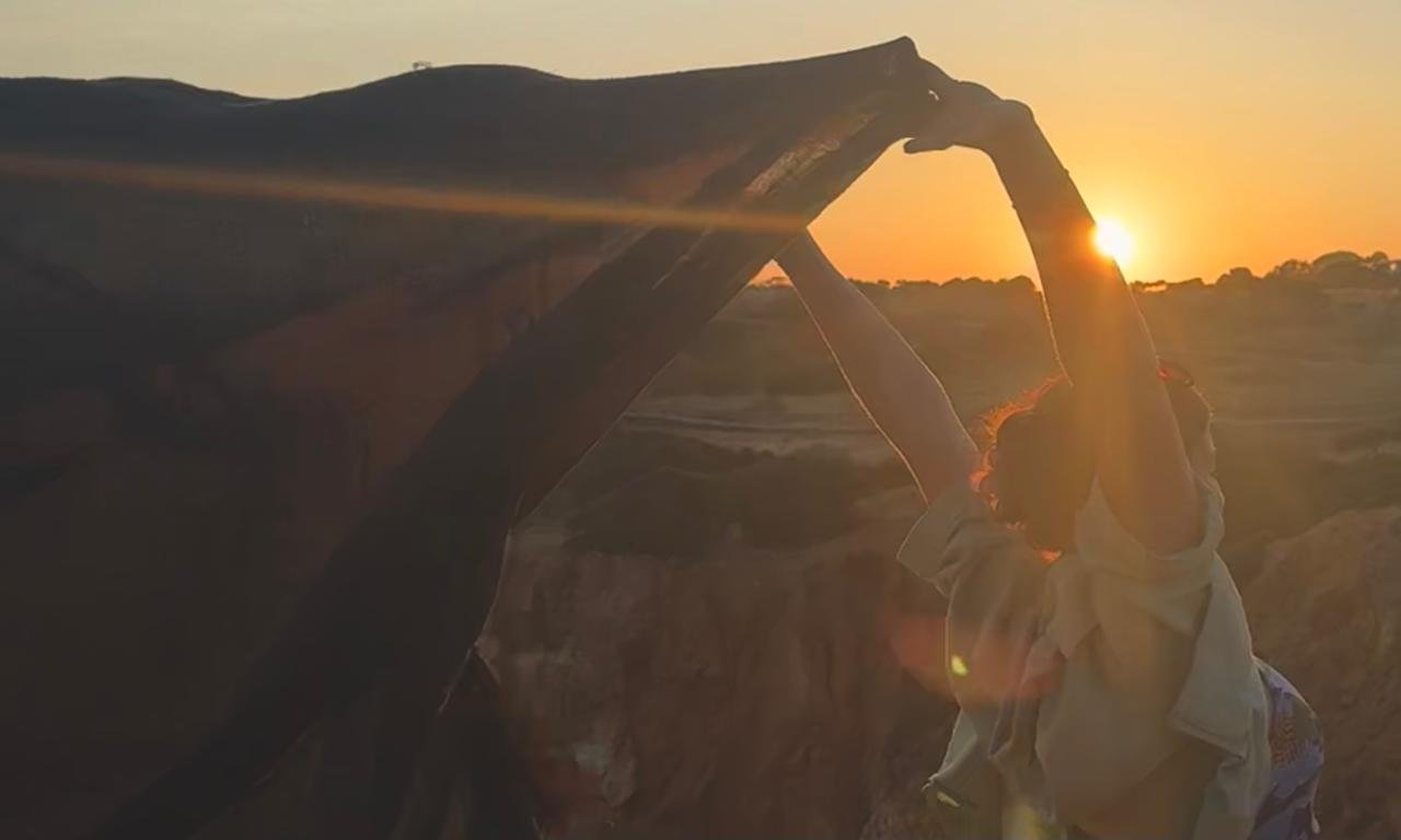 A person standing outdoors during sunset, holding a large stone arch with their arms raised. The setting sun is visible through the arch, casting a golden glow over the landscape.