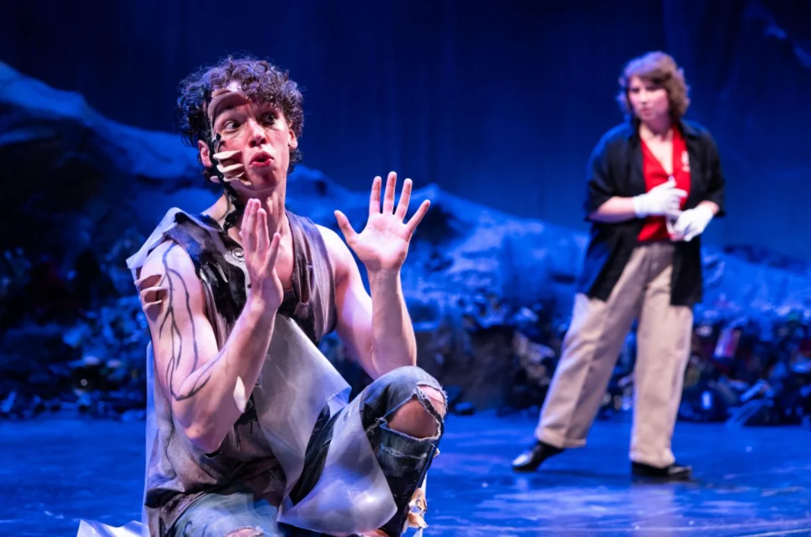 Two actors performing on stage; one actor in the foreground with curly hair, torn clothing, and expressive face, the other in the background with shoulder-length hair, wearing a black jacket and beige pants.
