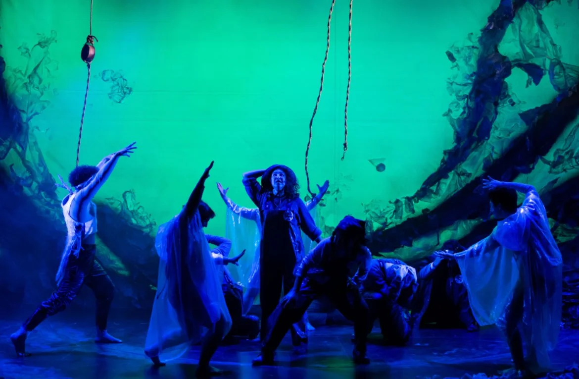A theatrical dance performance with seven dancers on stage, with a vibrant green and blue background, and hanging ropes. The dancers are in various dynamic poses, some reaching upward, others bent or crouched.