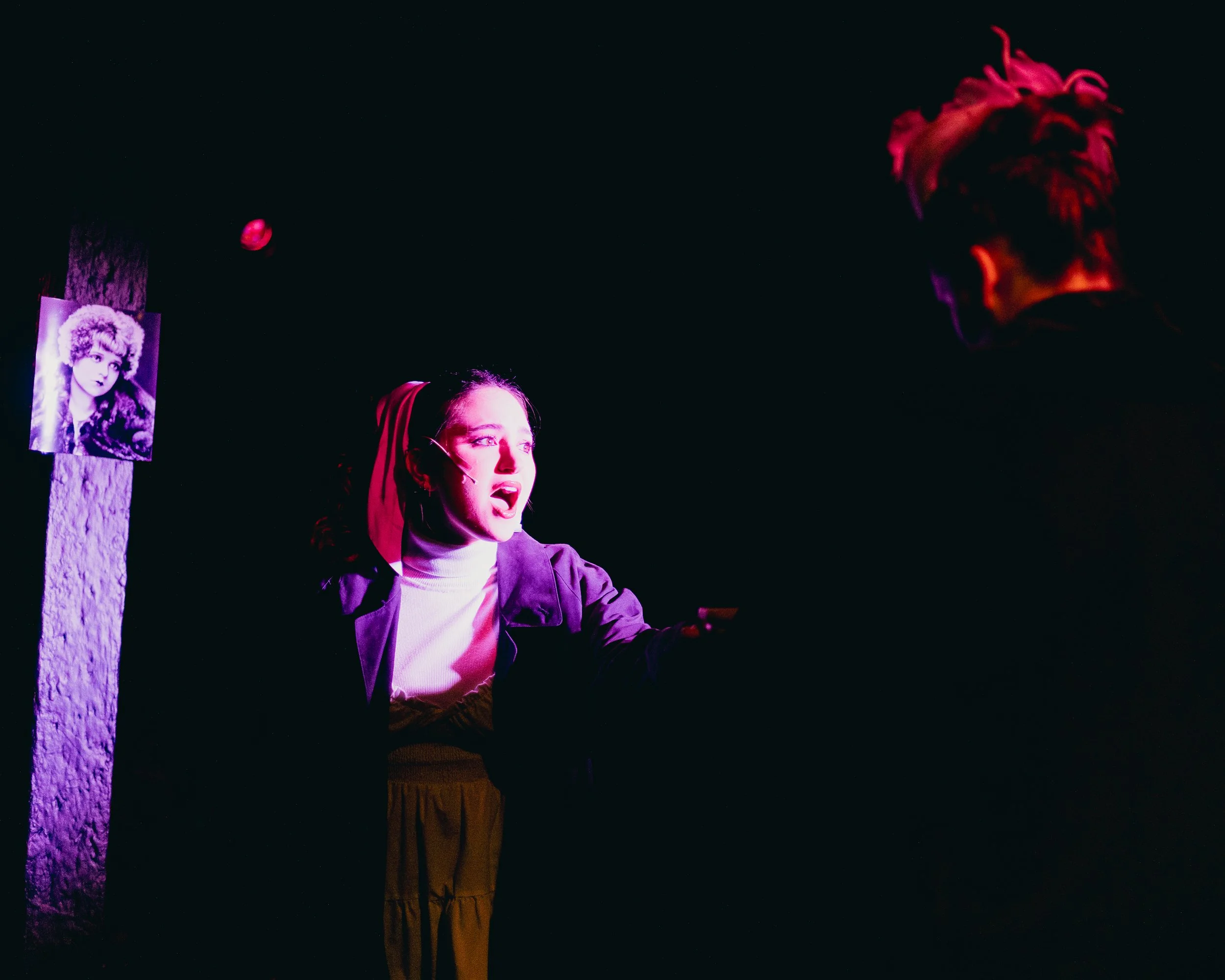 A theatrical scene with a woman singing or speaking passionately, illuminated by pink and purple stage lighting, with a man with pink hair listening, against a mostly dark background. A black and white portrait is visible on the wall to the left.