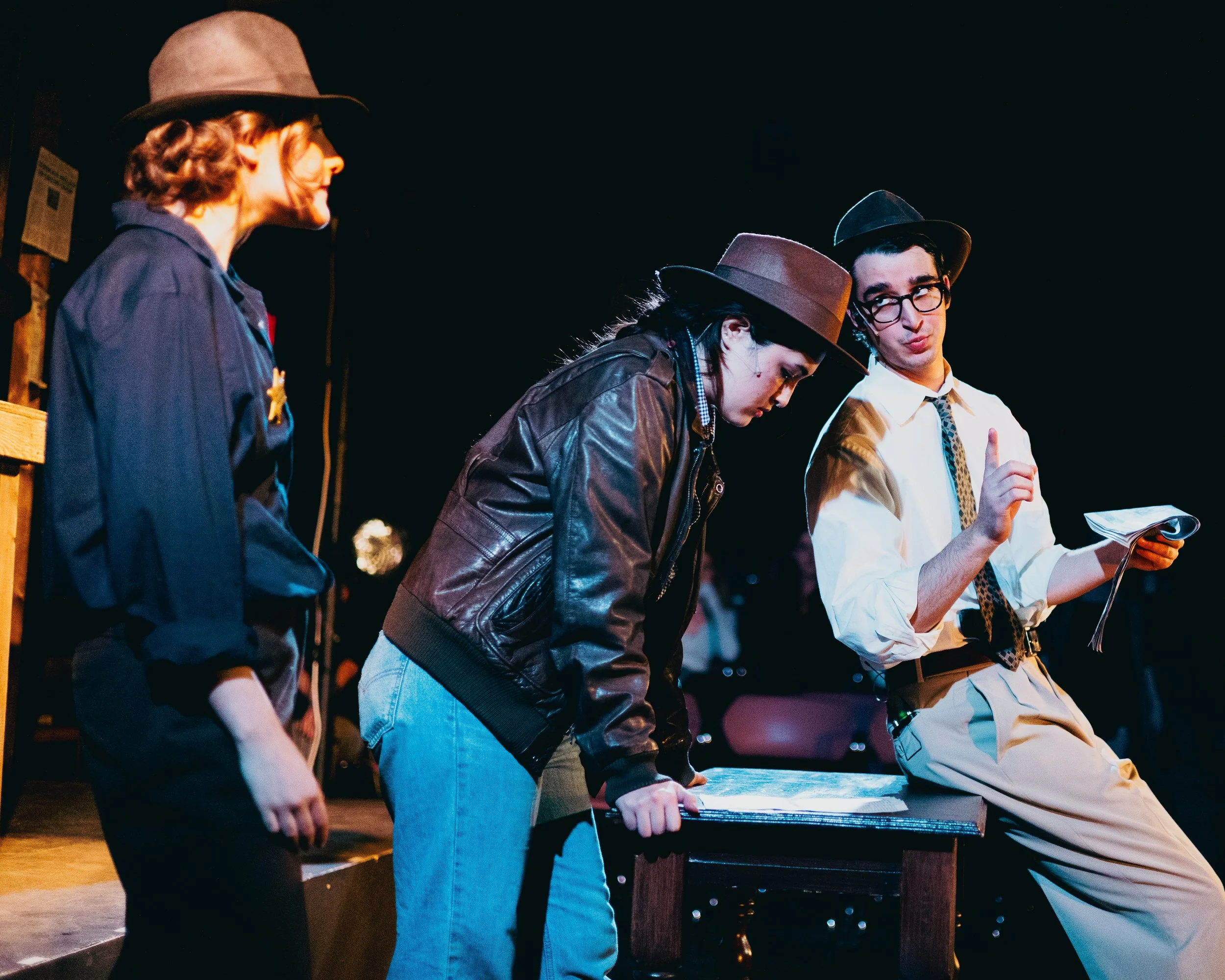 Three actors performing on stage in costume, with two men wearing fedoras and one woman in casual vintage attire, engaged in dialogue or scene.