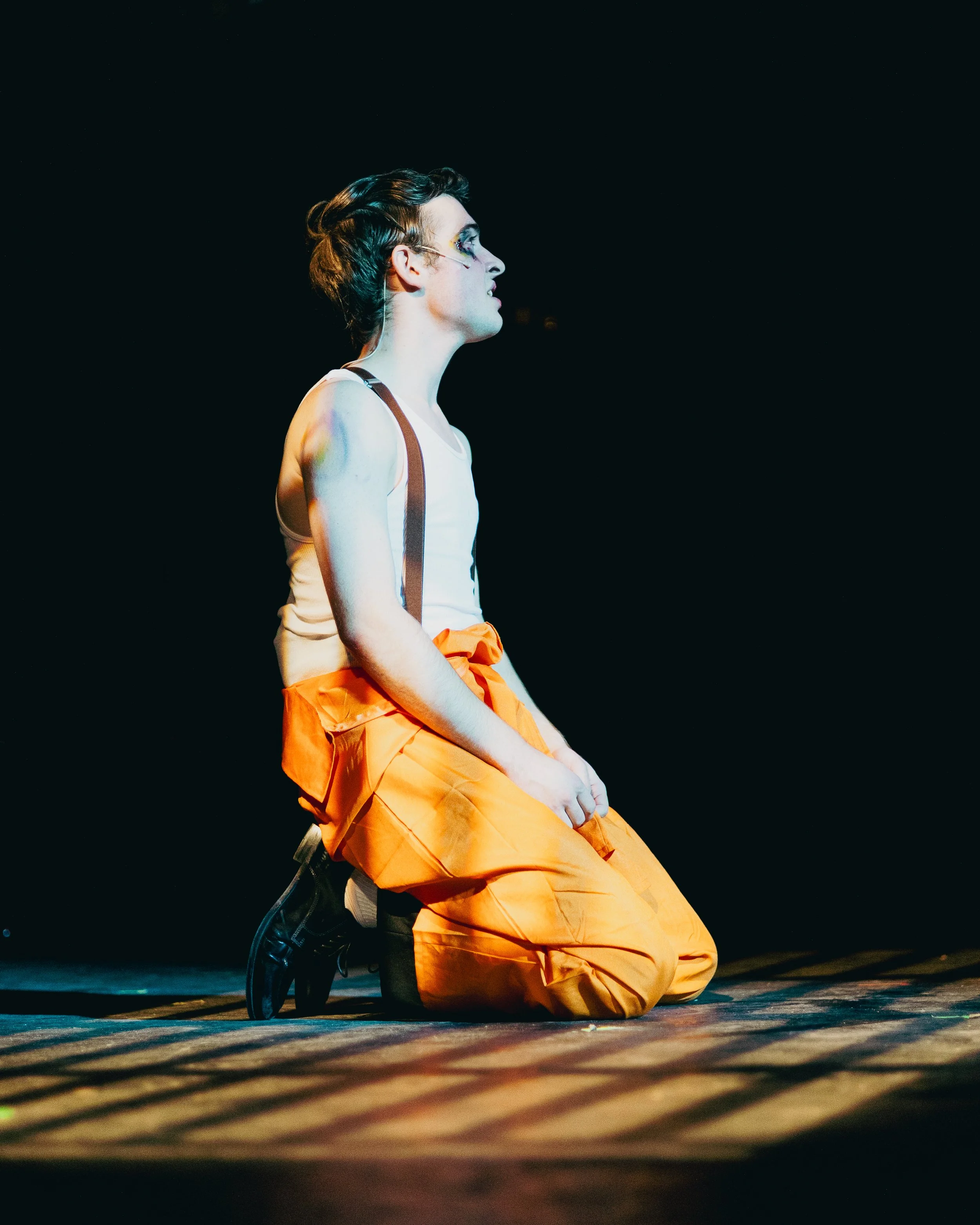 Person with makeup and glasses kneeling on stage, wearing orange pants, white tank top, and black shoes, against a dark background.