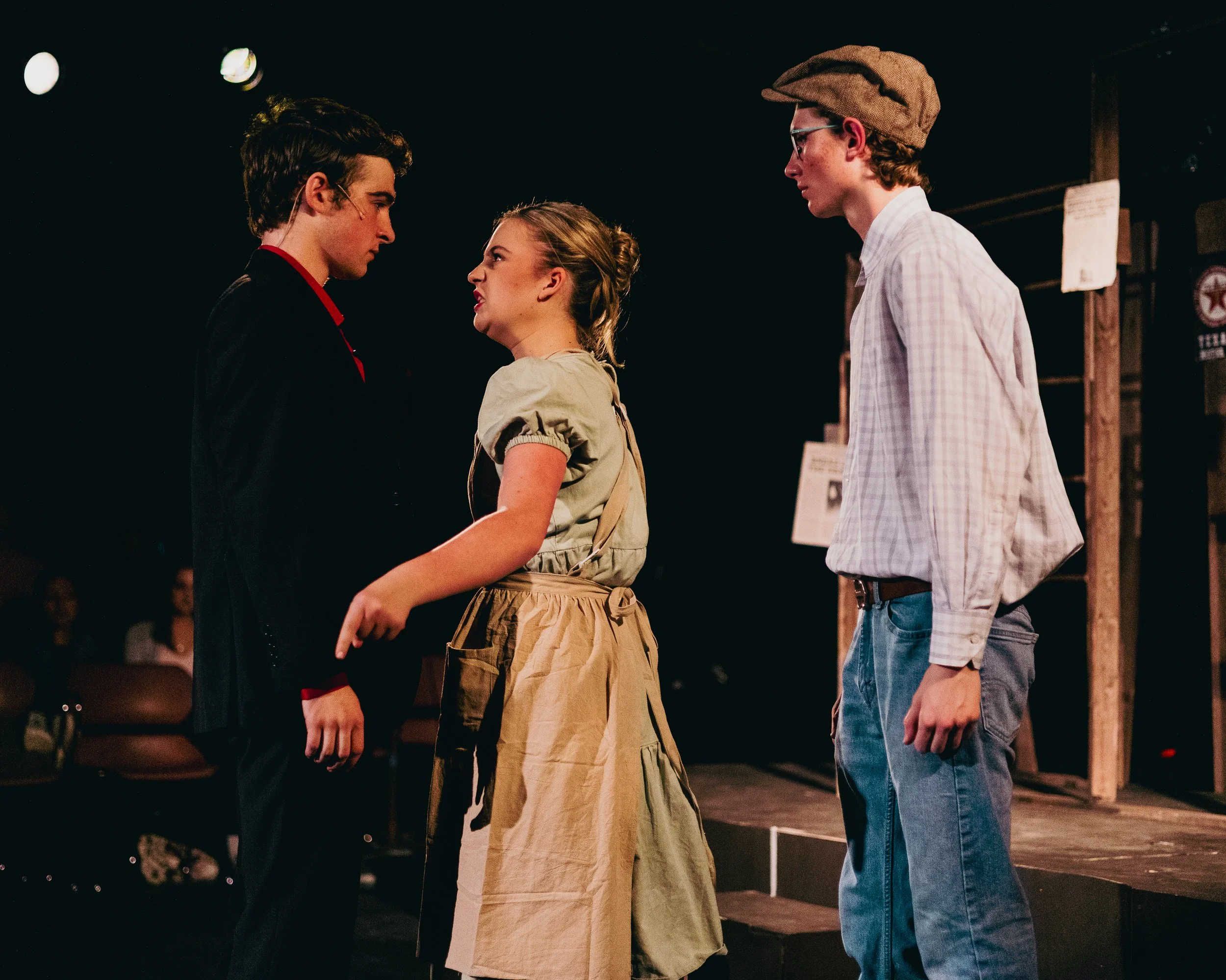 Three young actors on stage performing a scene with intense expressions. Two males and one female stand close, facing each other. The backdrop is dark with stage lights.