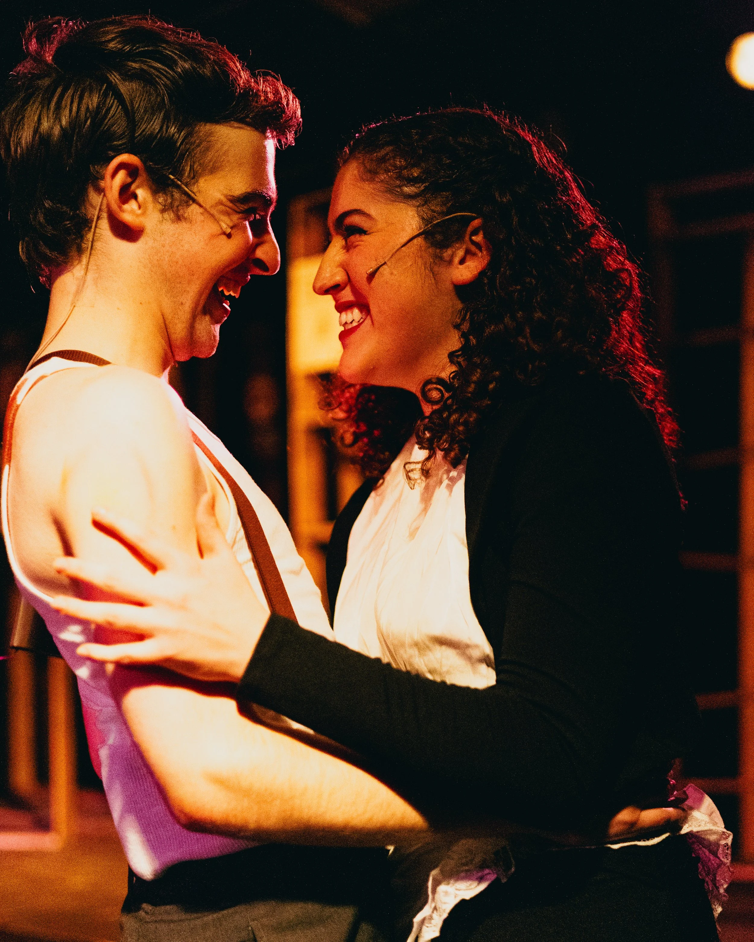A young man and woman are smiling and looking at each other while dancing closely together in a warmly lit indoor setting.