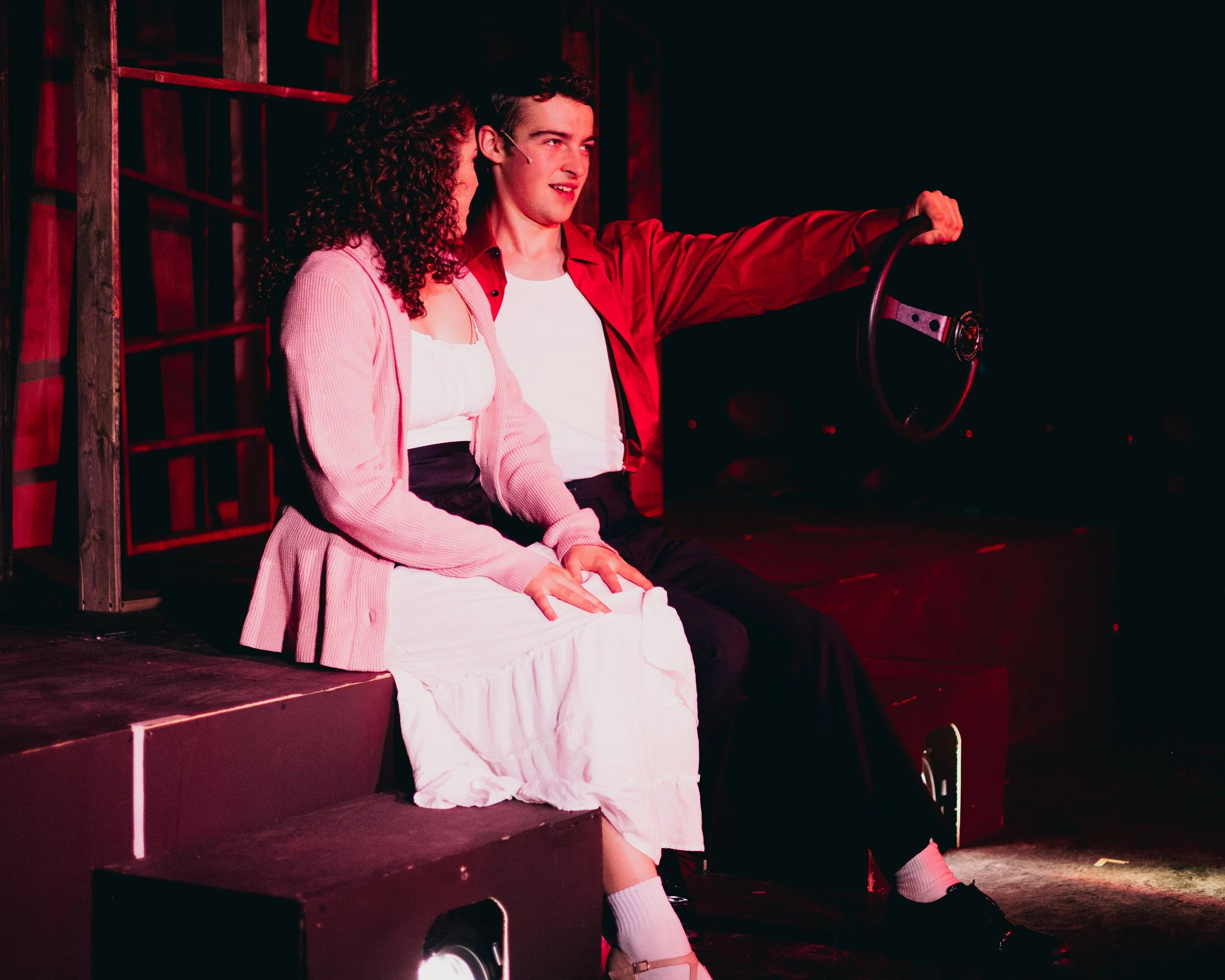 Two young actors sit on a stage with a dark background. The male actor, dressed in a red jacket and white shirt, holds a steering wheel prop, while the female actor in a pink cardigan and white dress sits beside him. The scene is lit with red stage l