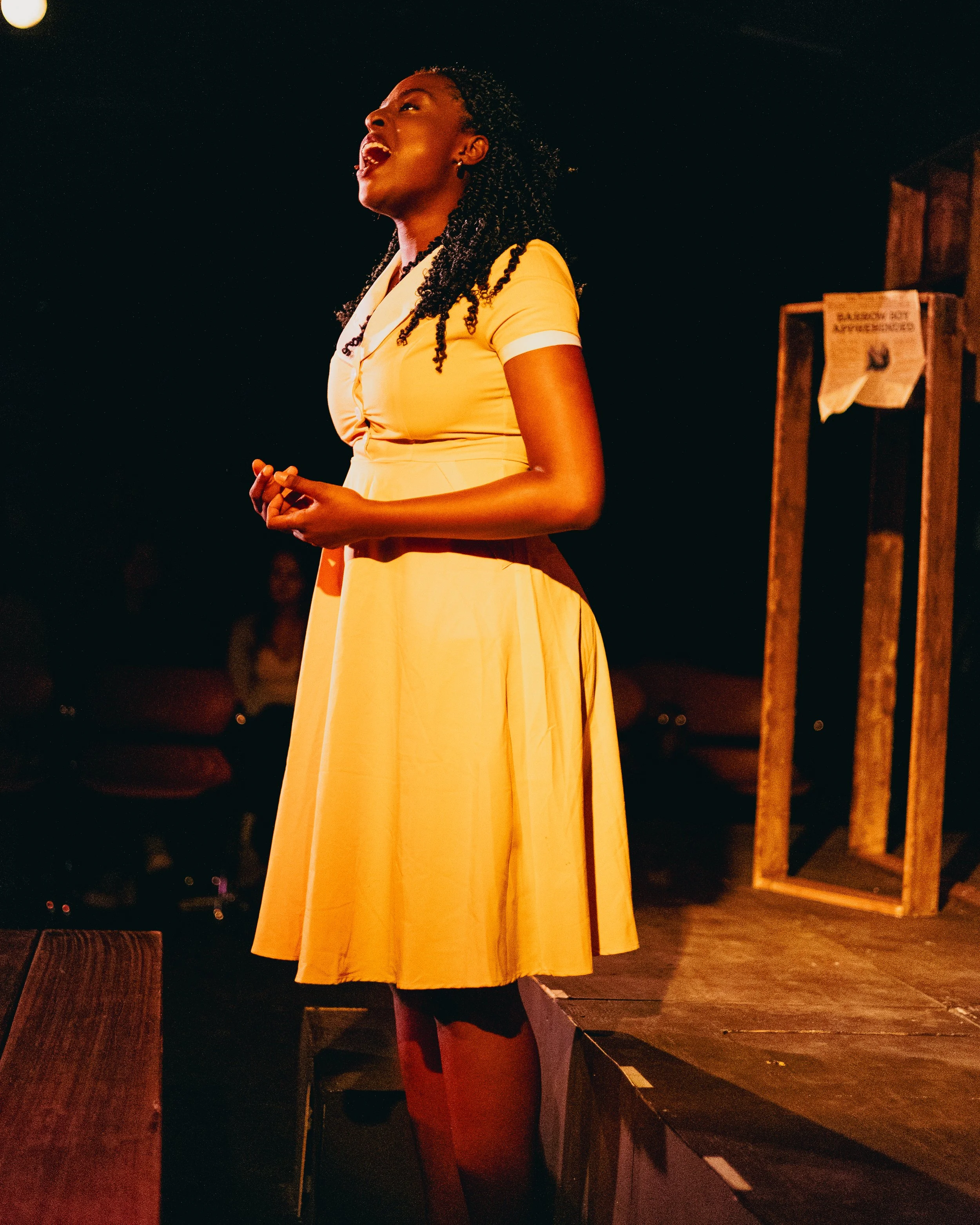 A woman in a yellow dress standing on a stage, speaking or singing passionately, with a dark background and a wooden frame behind her.