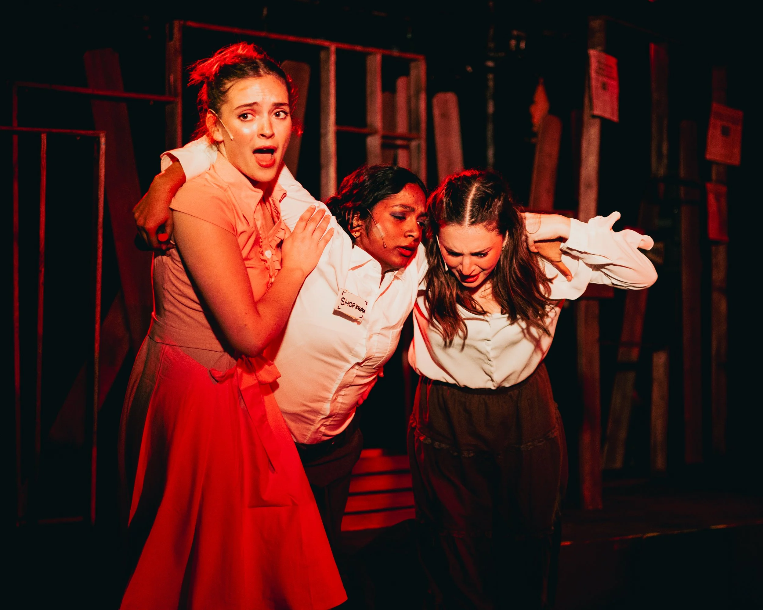 Three women on stage acting out an emotional scene, with one woman in a red skirt and two women in white shirts, holding each other with distressed expressions.