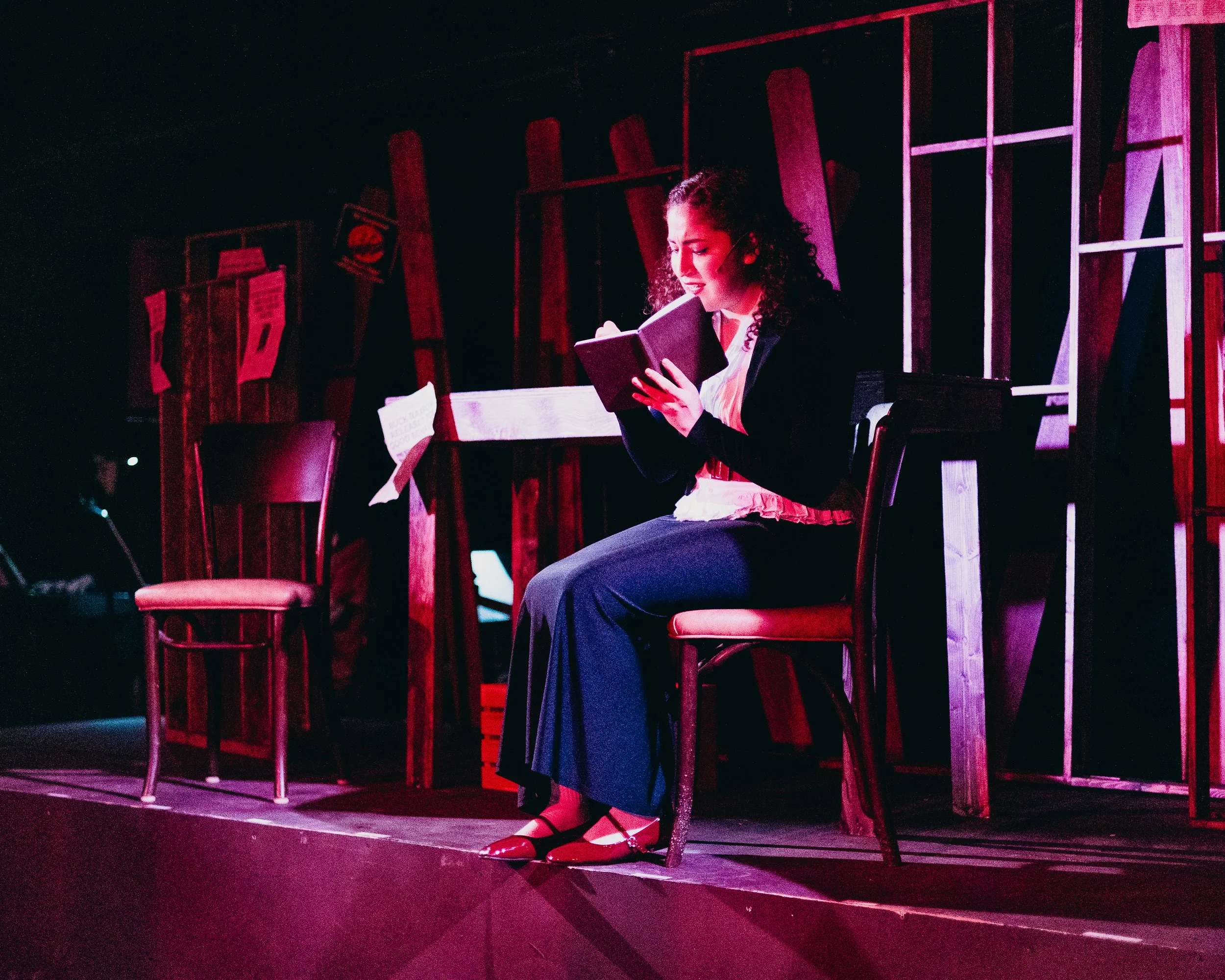 A woman sitting on a chair on stage, reading a book, with a dimly lit background featuring a man in the background and a wooden wall with papers pinned to it.