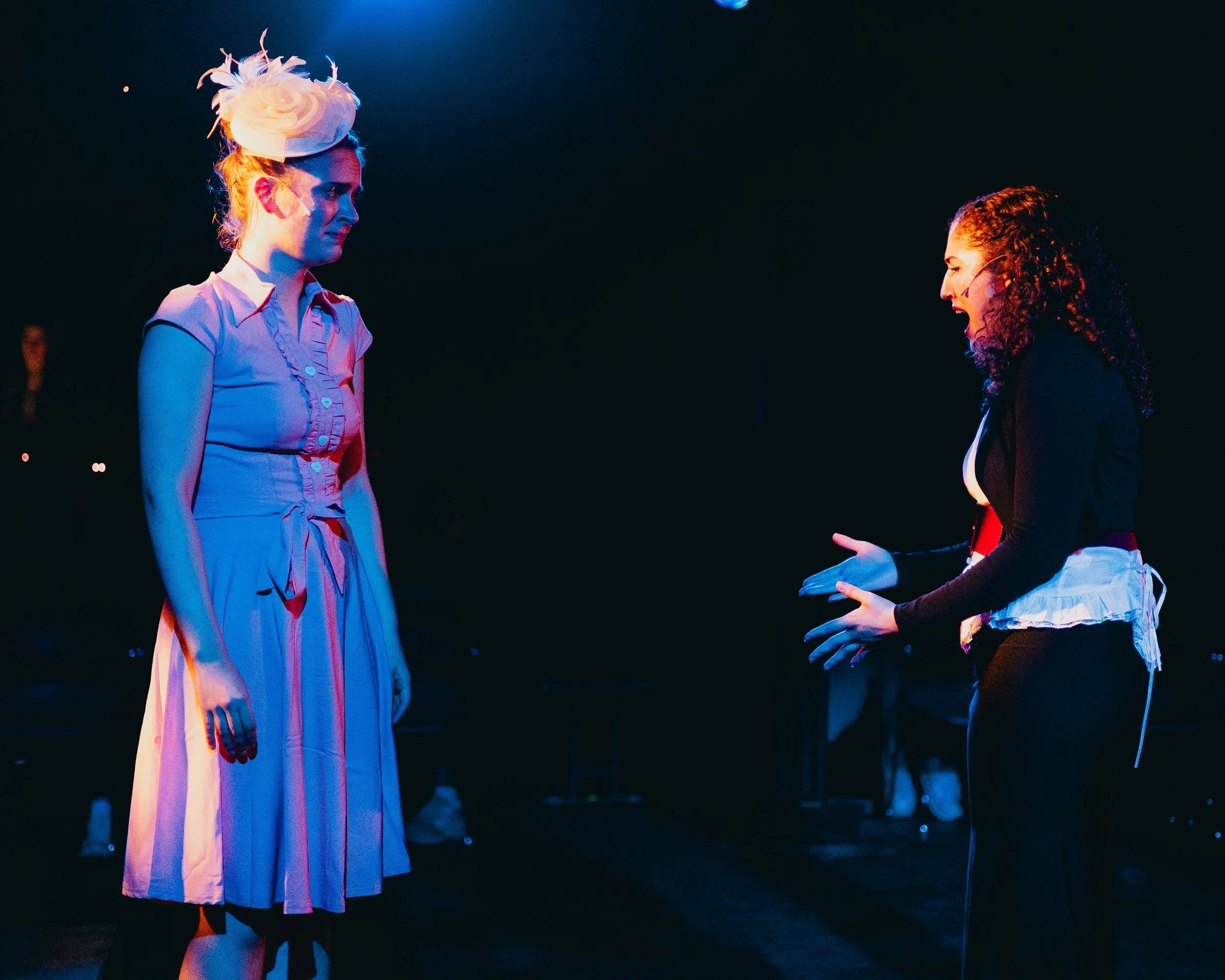 Two women on stage, one with dark curly hair and a headset microphone, speaking passionately, and the other with light hair and an elaborate hat, standing quietly, in the spotlight.