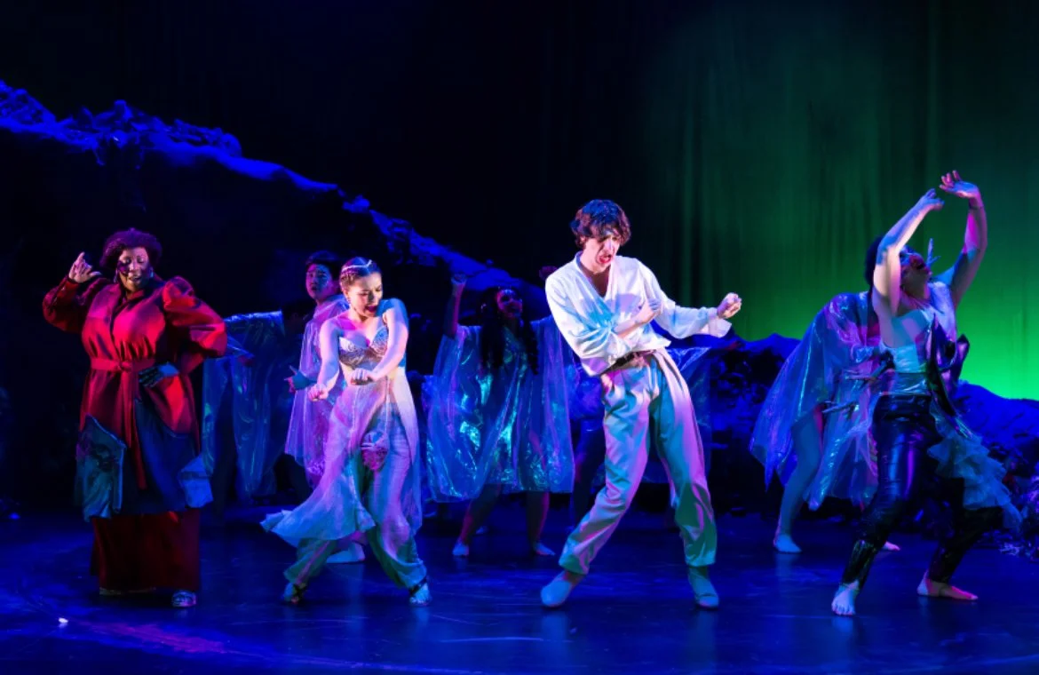 A group of dancers performing on stage with colorful costumes, blue and green stage lighting, and a dark scenic background.