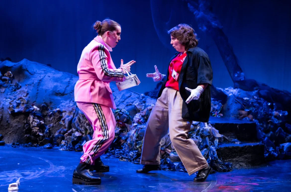 Two women dressed casually confronting each other on stage with a backdrop of a trash heap and dark blue lighting.