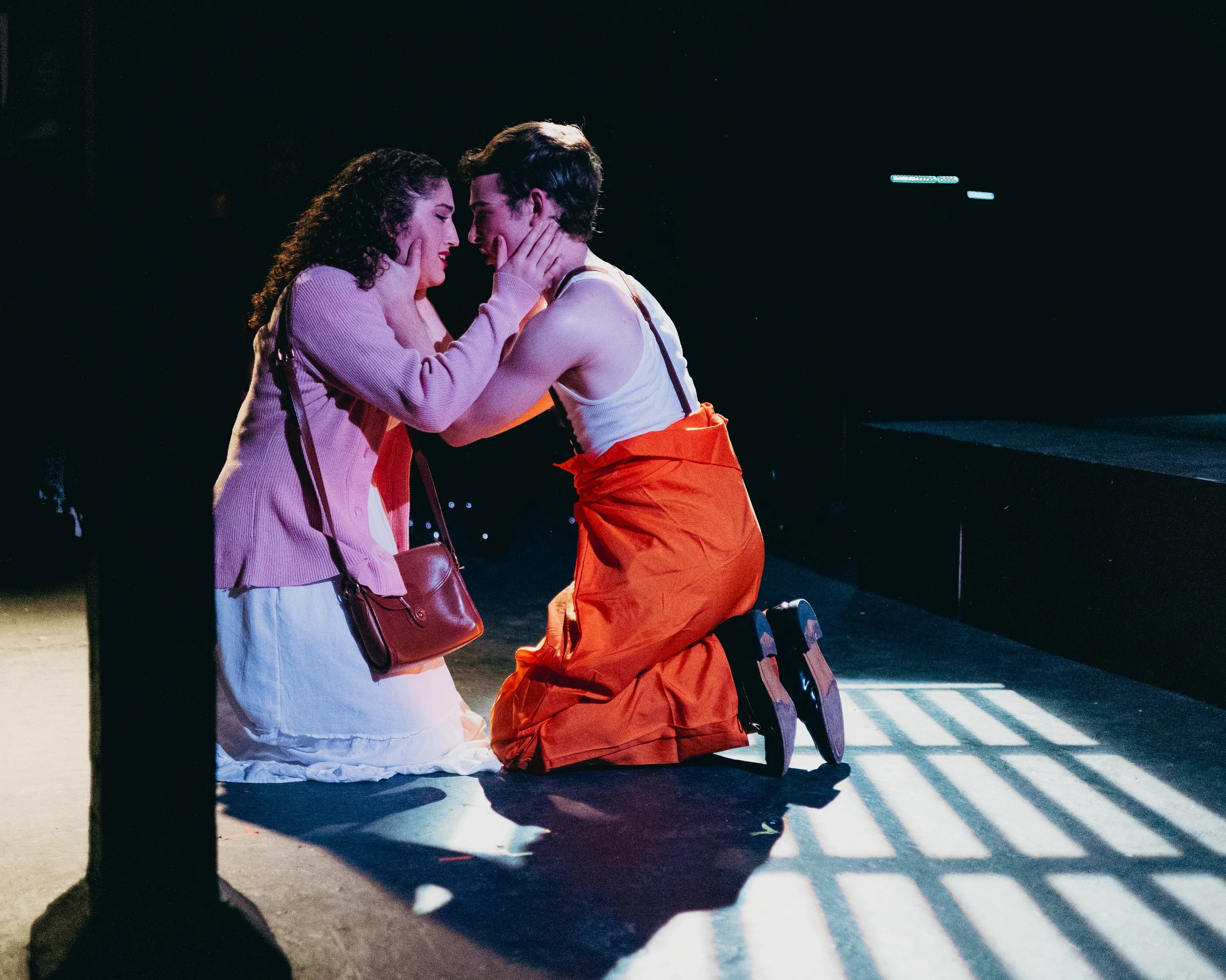 Two women on stage, one in a pink jacket and white skirt, the other in an orange skirt and white tank top, kneeling and touching faces while holding each other's faces, with dramatic lighting creating shadows.