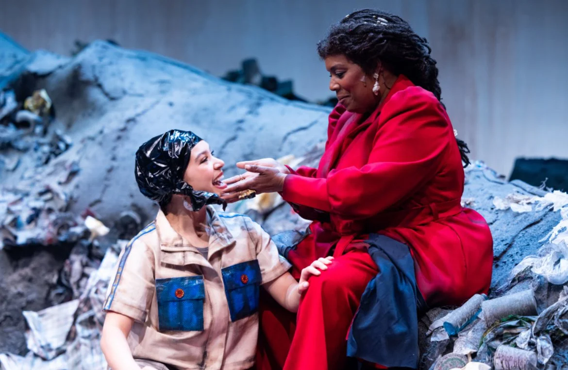 Two women sitting amidst trash, one wearing a beige outfit and a black plastic cap, the other in a red suit, smiling at each other.
