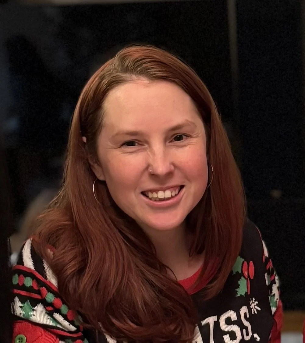 A woman with red hair smiling at the camera, wearing a Christmas sweater with holiday patterns.