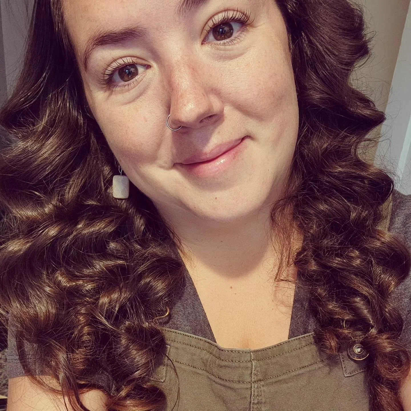 Close-up of a woman with brown curly hair, wearing earrings and a nose ring, smiling at the camera.