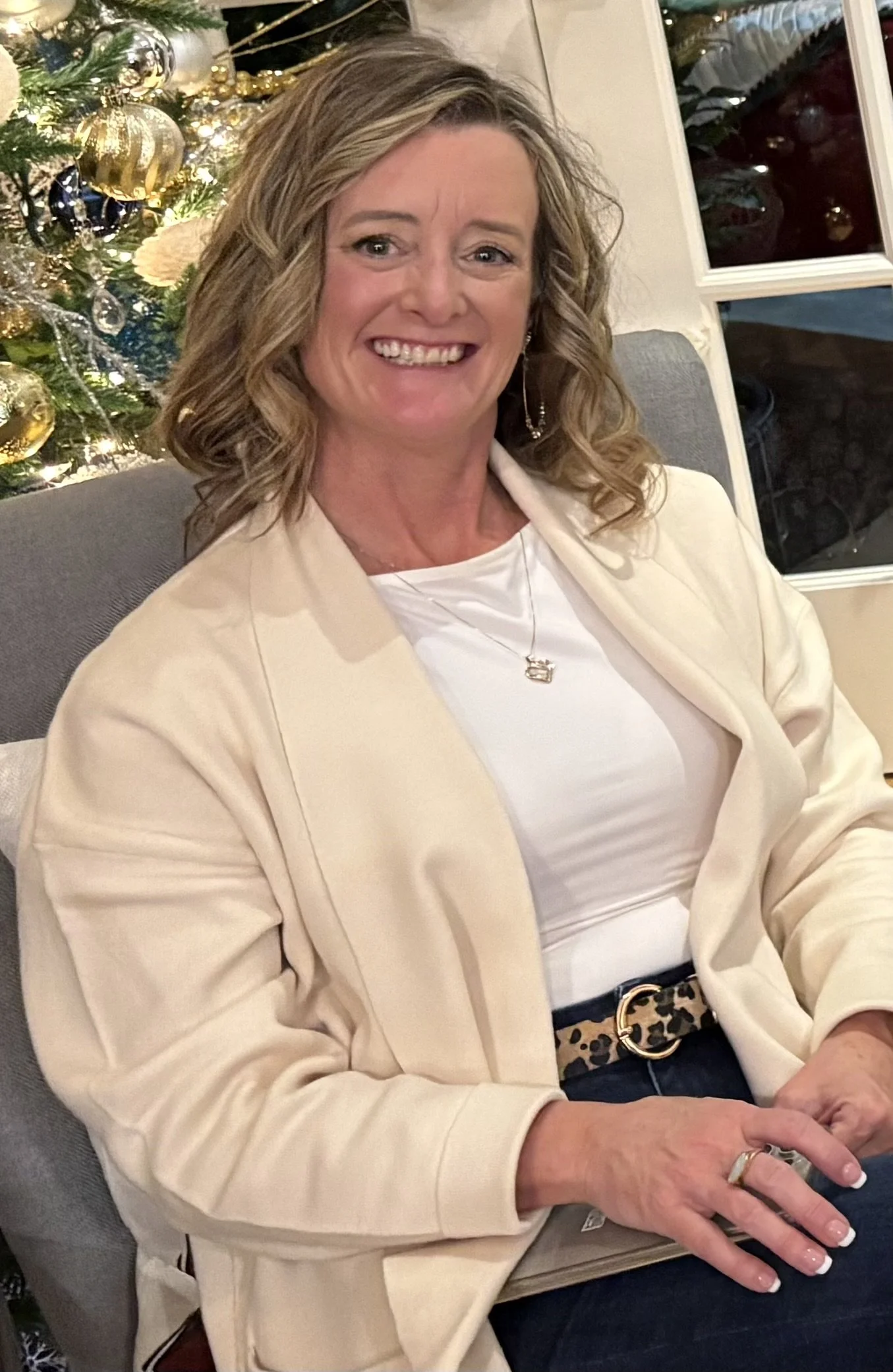 A woman with curly blonde hair smiling, seated near a decorated Christmas tree with gold and silver ornaments, wearing a white top, beige blazer, dark jeans, and a patterned belt.