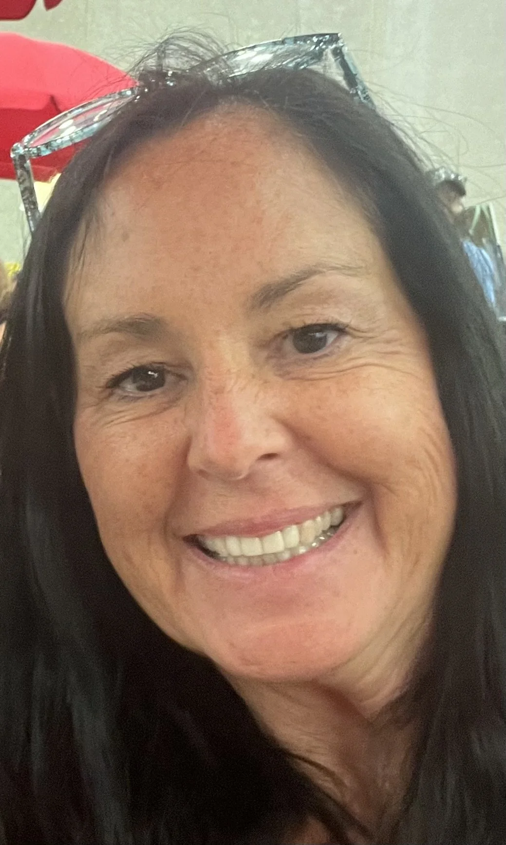 A close-up of a smiling woman with dark hair and sunglasses on her head, in an indoor setting with a red umbrella in the background.
