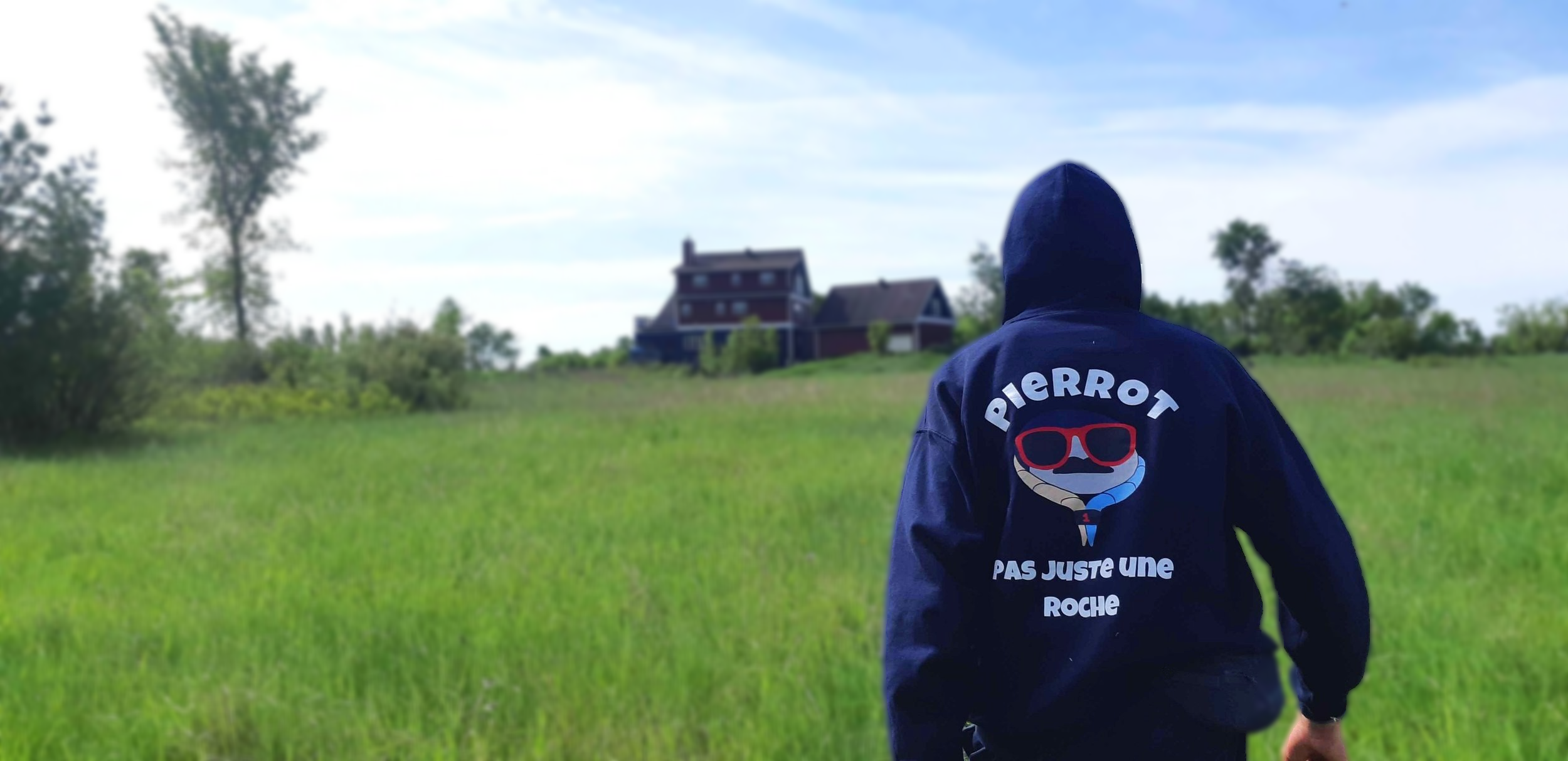 Person portant une veste avec le texte 'Pierrot, Pas Juste une Roche' marche dans un champ vert vers une maison en arrière-plan sous un ciel bleu avec quelques nuages.
