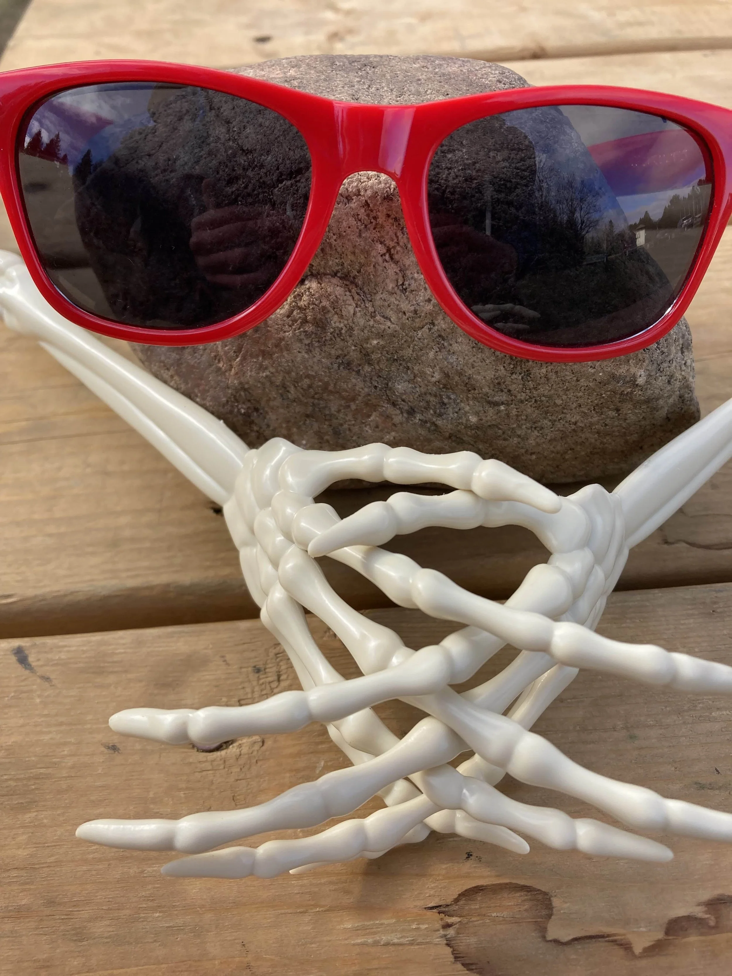 Lunettes de soleil rouges reposant sur une pierre, avec une structure blanche imitant des branches ou des os en dessous, posées sur une surface en bois.