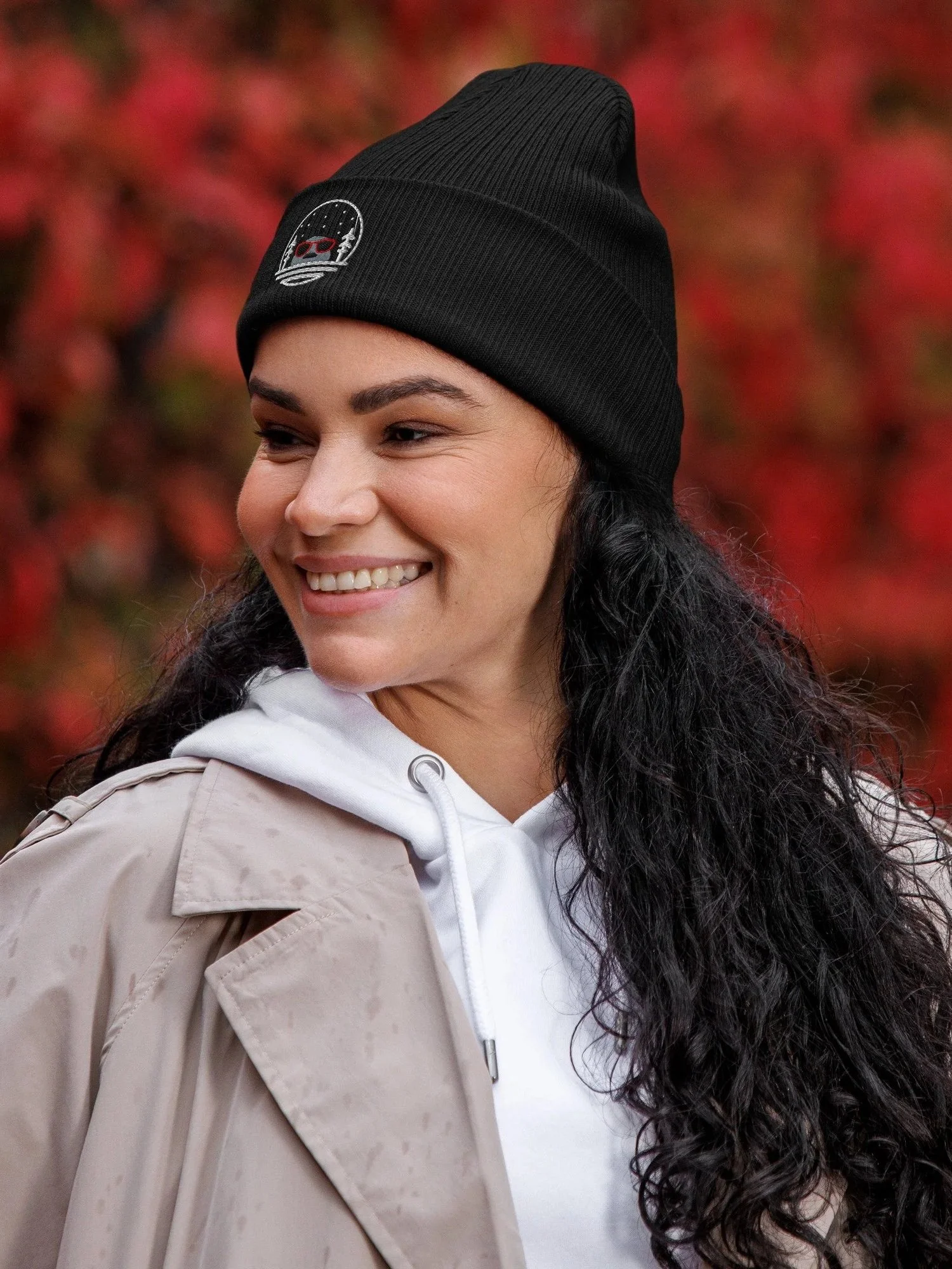 Femme souriante portant un bonnet noir et un manteau beige, avec un fond de feuilles rouges d'automne.