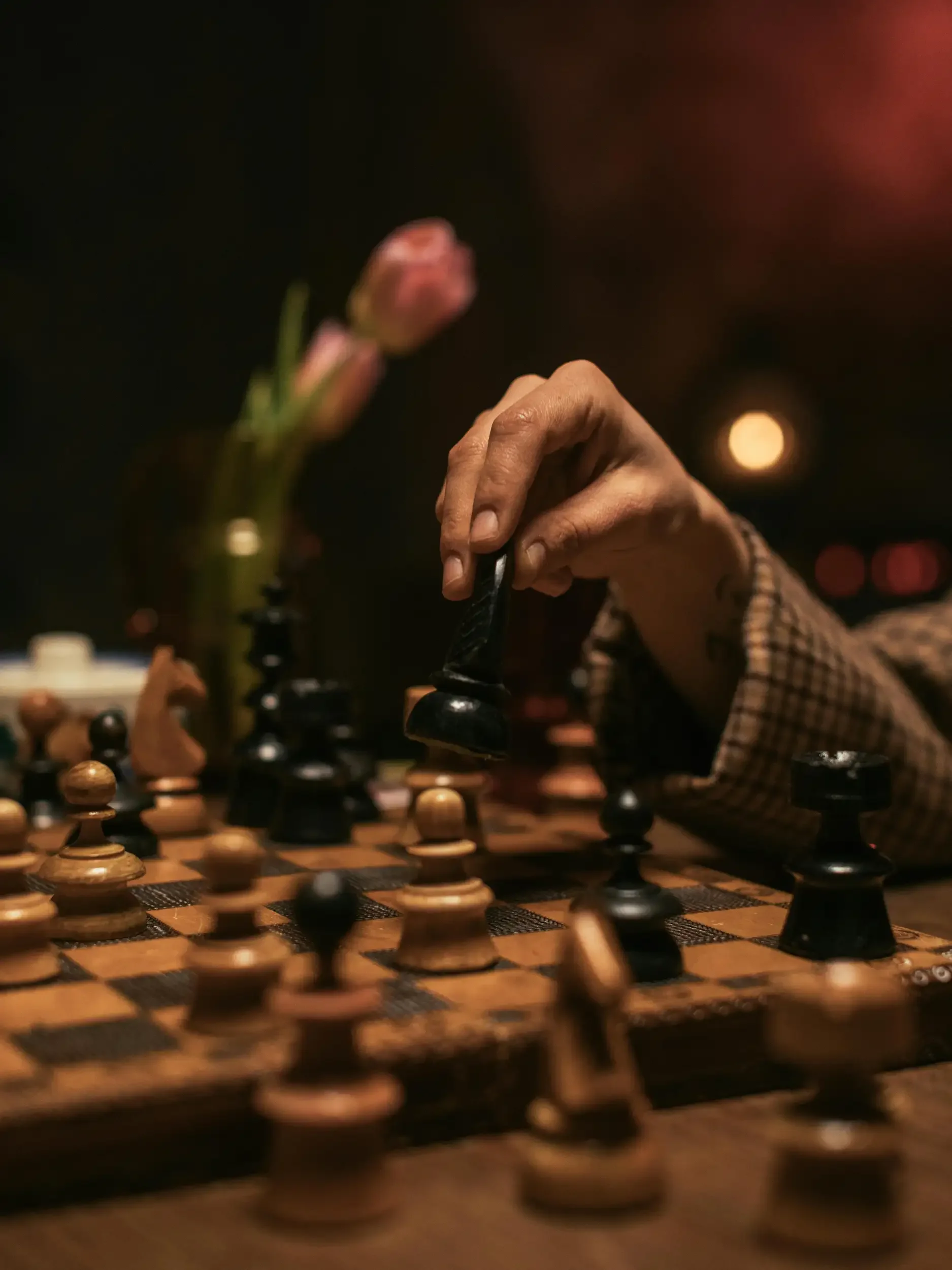 Close-up of a person's hand making a move in a chess game on a wooden chessboard, with a blurred background and pink flowers in a vase.