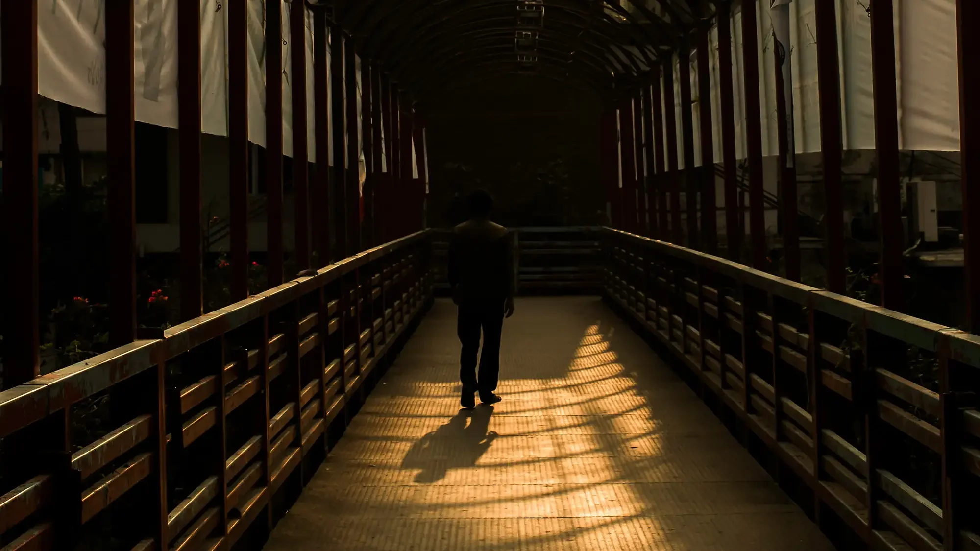 A person walking on a bridge at sunset, with long shadows cast by the bridge's railing and structure.