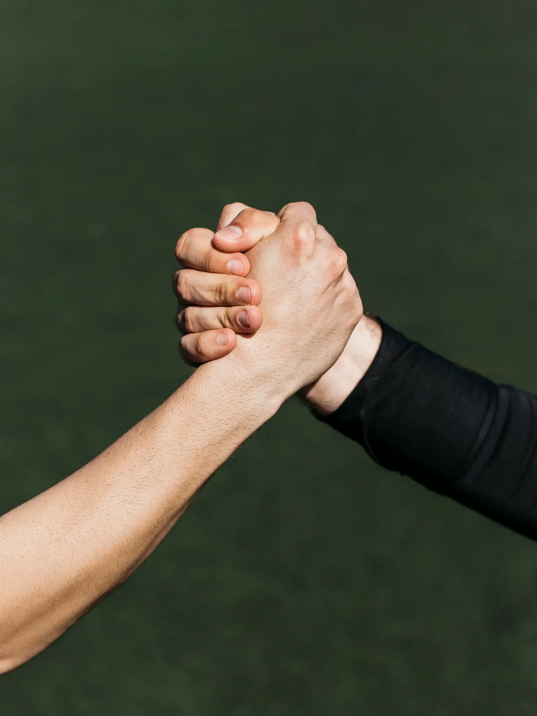 Two people clasp their hands in a handshake or fist bump, one wearing a black sleeve, against a green background.
