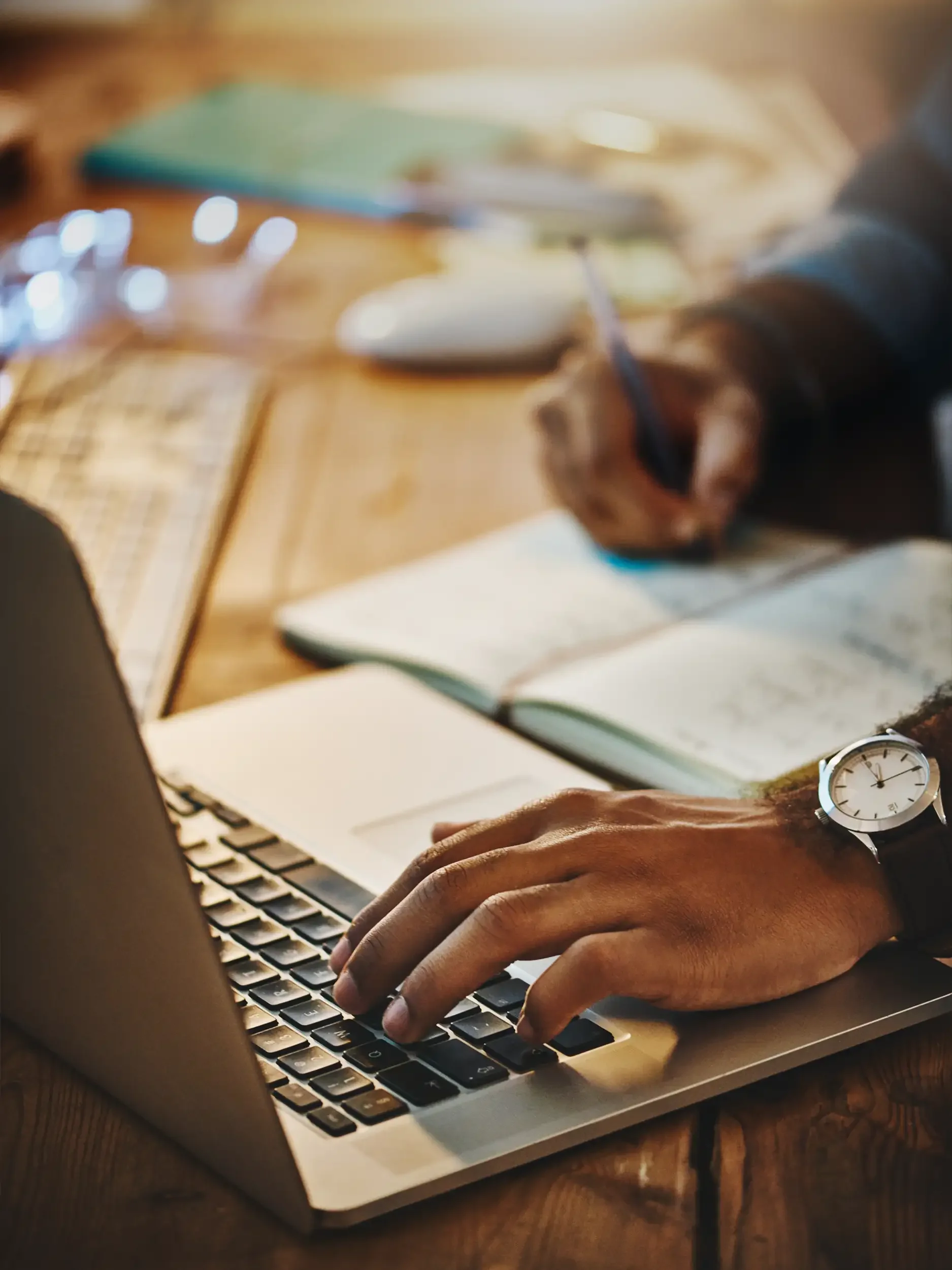 Close-up of a person's hand typing on a laptop keyboard at a wooden desk, with notebooks, a mouse, and glasses in the background.