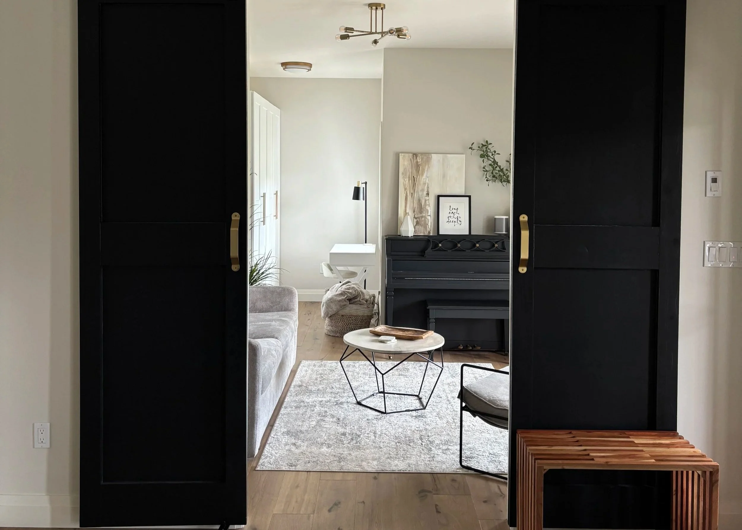 Living room view through black sliding closet doors, showcasing a gray sofa, white side table, black musical keyboard, artwork, and decorative plants with natural light.