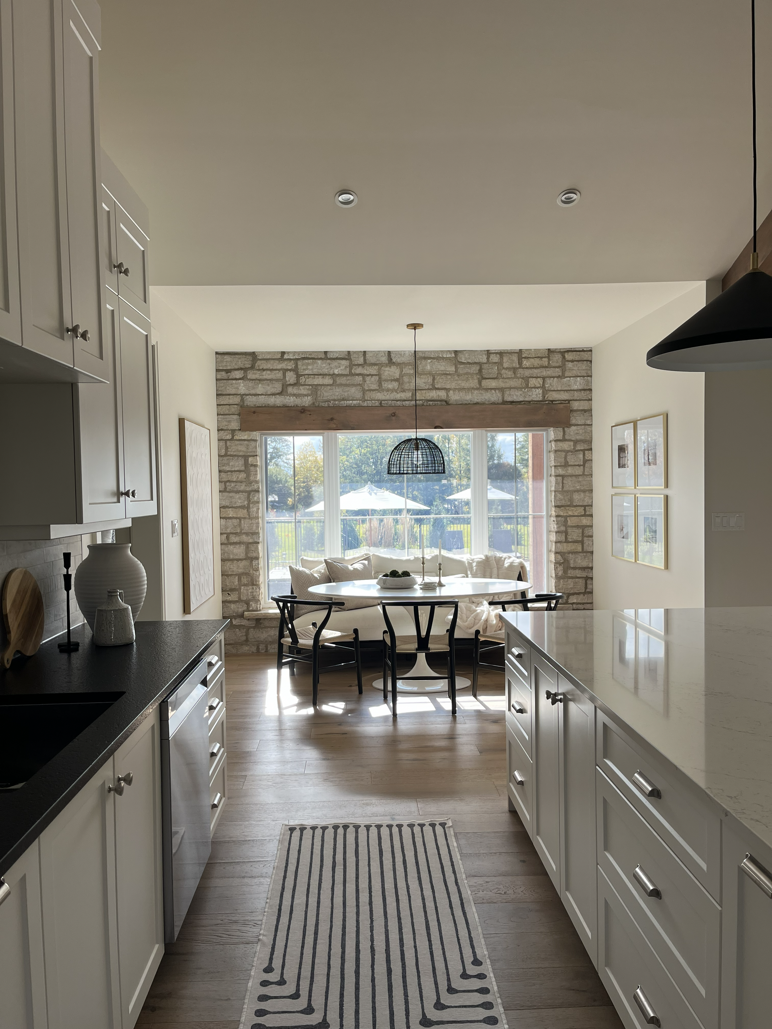 View of a bright kitchen with white cabinets, a black countertop, and a striped rug on wooden flooring. The background features a dining area with a round table, black chairs, and a window with a brick wall and outdoor scenery.