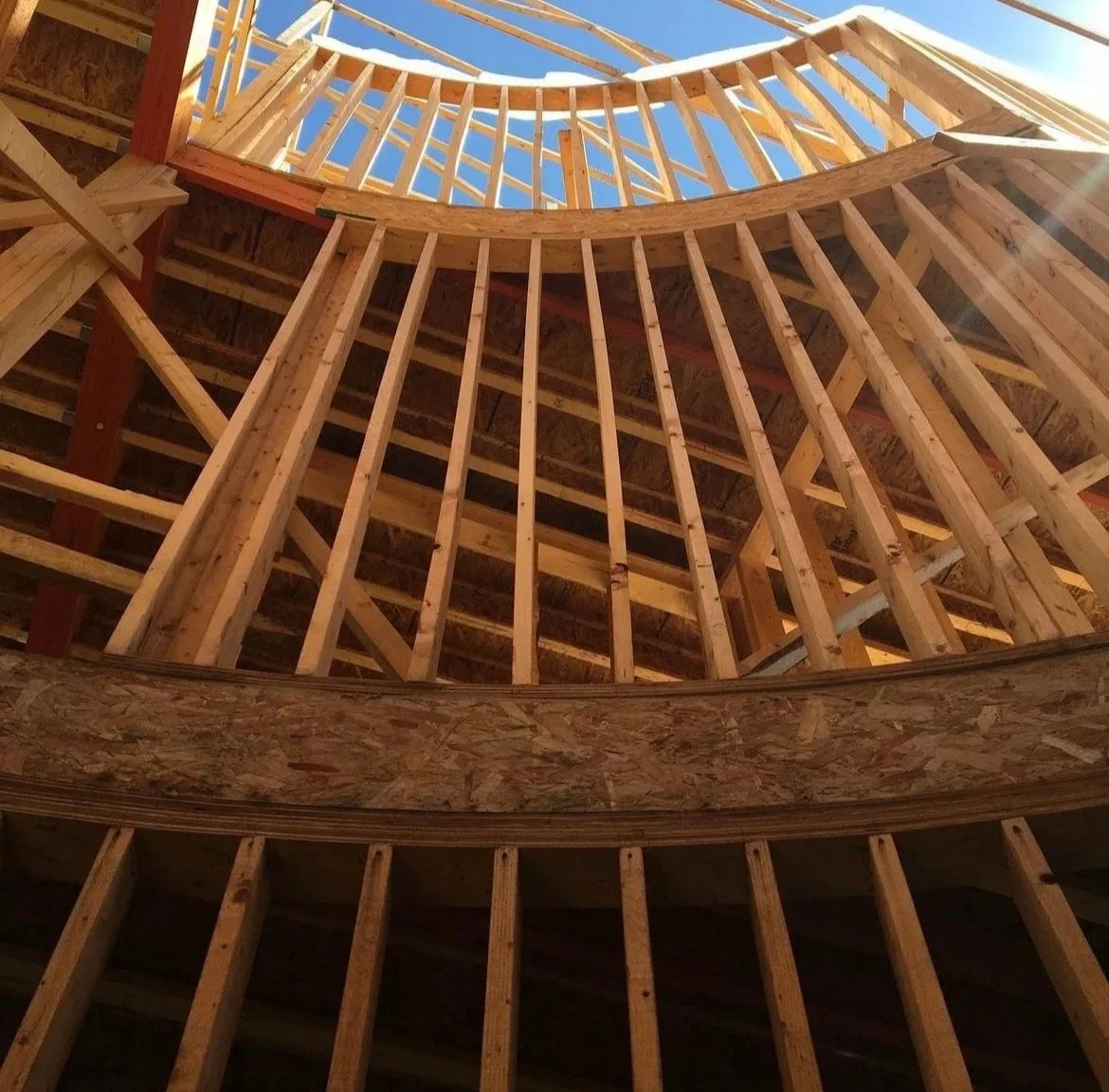 Looking up from inside a curved wooden building under construction, showing a circular opening at the top sky and wooden framing.