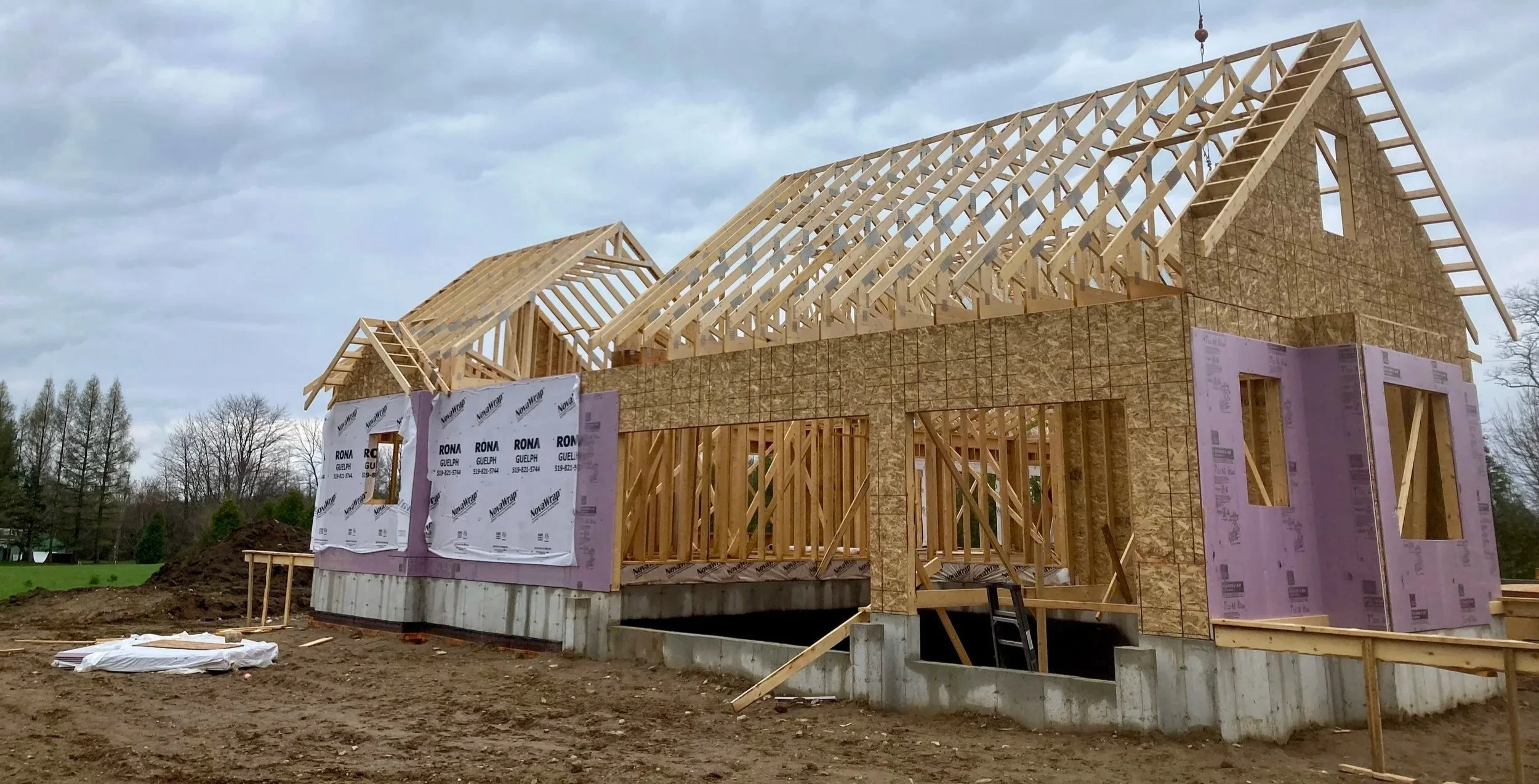 A house under construction with a wooden frame, oriented in a rectangular shape with a pitched roof. There are multiple window openings, and the walls are partially covered with plywood. The construction site has dirt and construction materials aroun