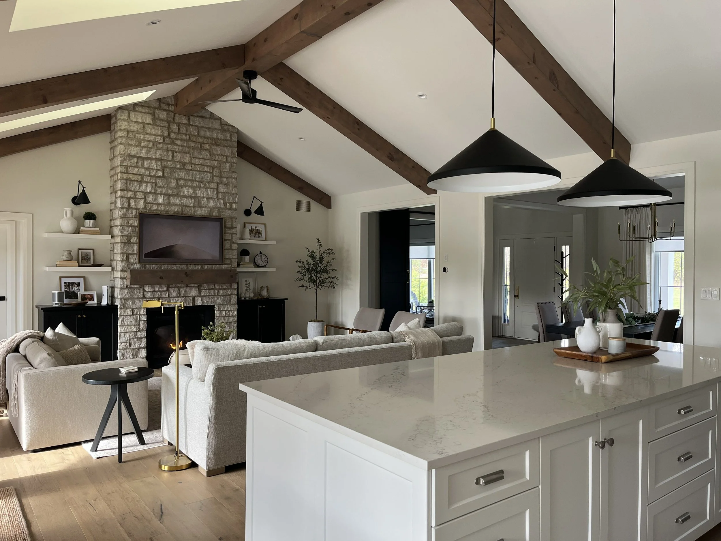 Open-concept living room with beige sofas, a stone fireplace, wooden beams on the ceiling, black and gold pendant lights, a black sideboard, and decorative shelves with plants and photos.