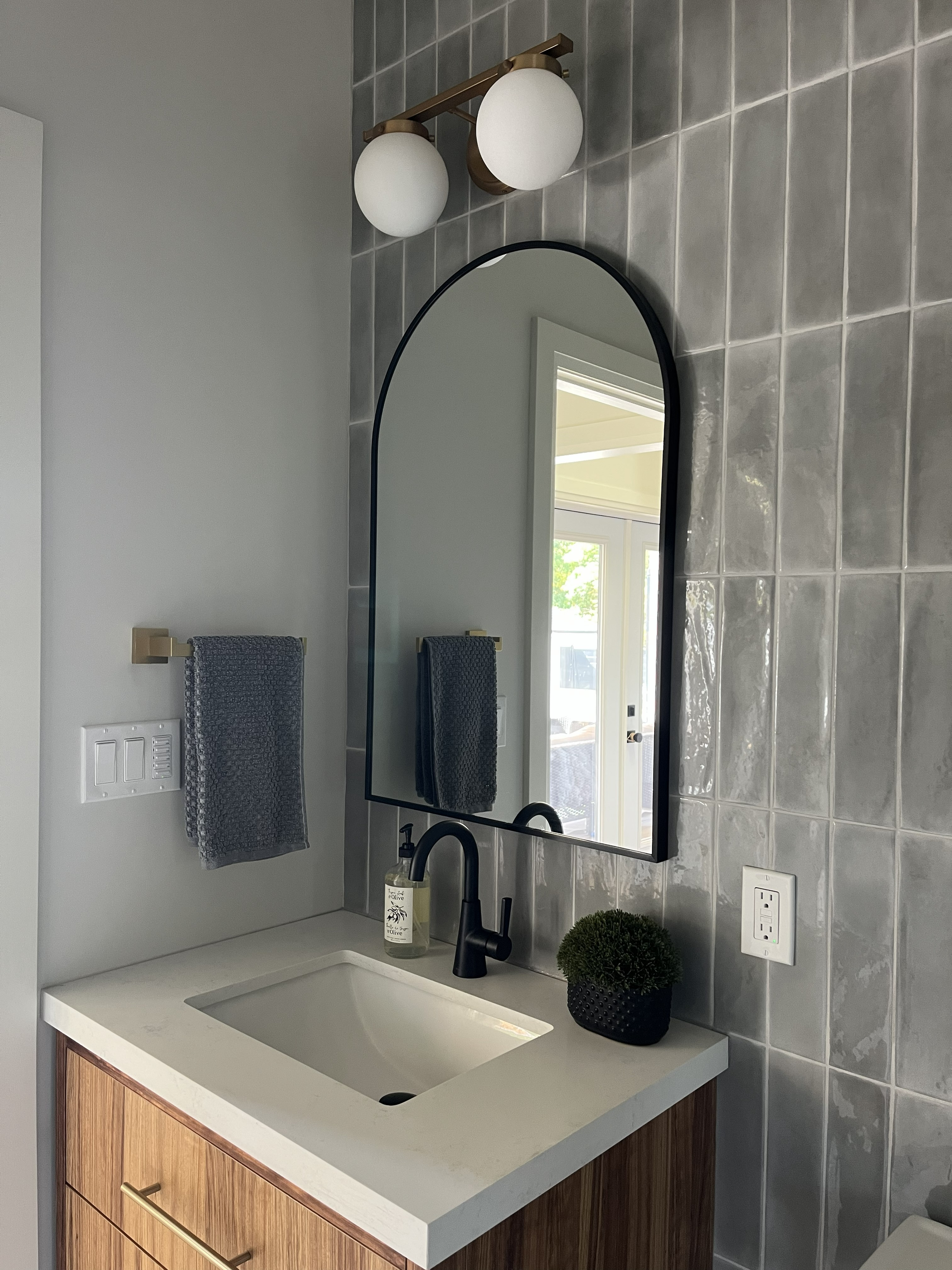 Bathroom vanity with wooden cabinet, white sink, black faucet, mirror, two gray hand towels, soap dispenser, and a potted green plant, with gray tiled wall and window.
