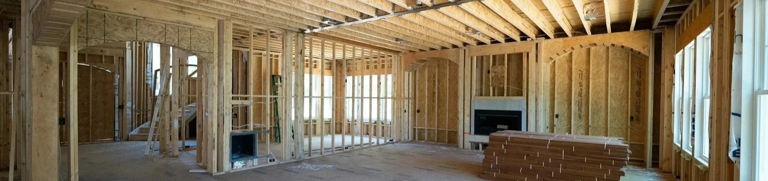 Interior view of a house under construction with exposed wooden framing and insulation, a new fireplace on the wall, and large windows along the right side.